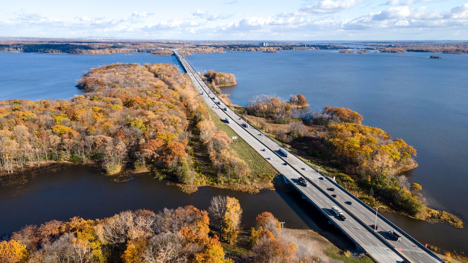 Aerial view of a long highway bridge crossing a vast lake surrounded by colorful autumn foliage trees on a sunny day.