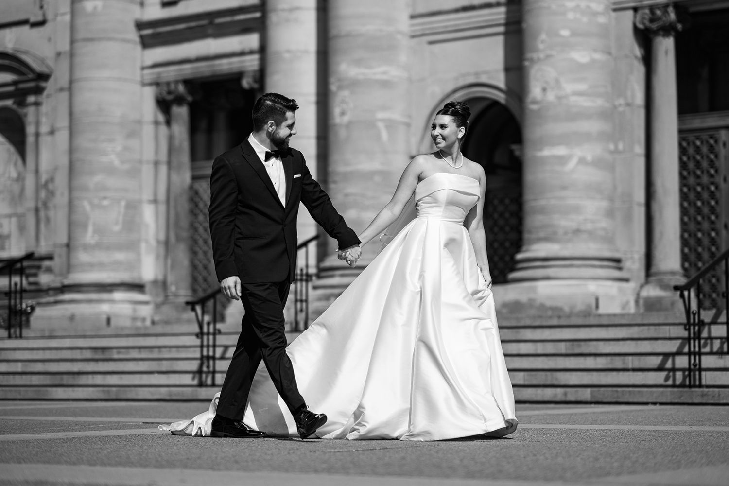 Black and white photo of a bride and groom holding hands walking in front of a grand building on their wedding day.
