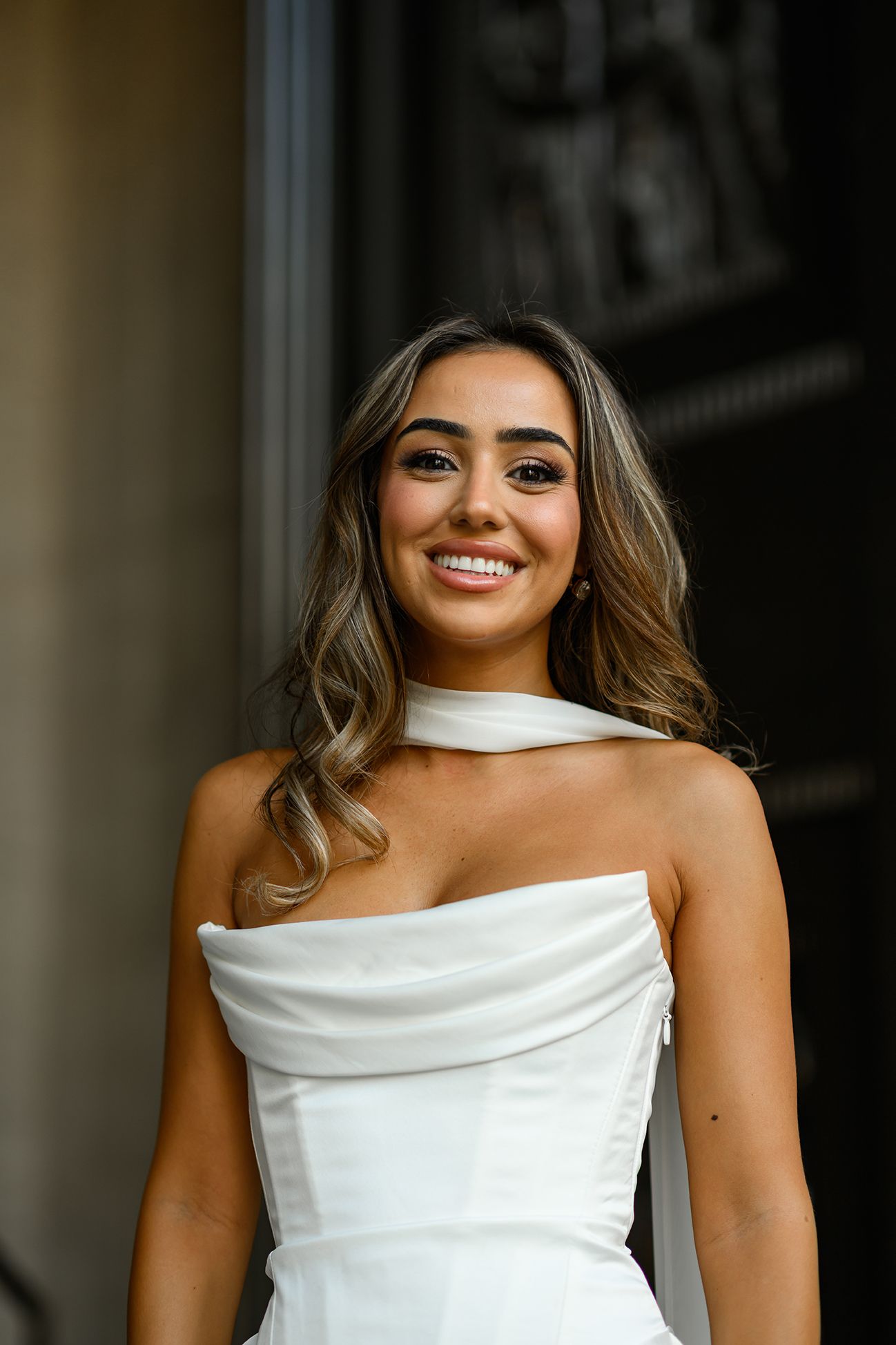 Smiling woman in white strapless gown with choker neckline, long wavy hair, posing outdoors near elegant columns.