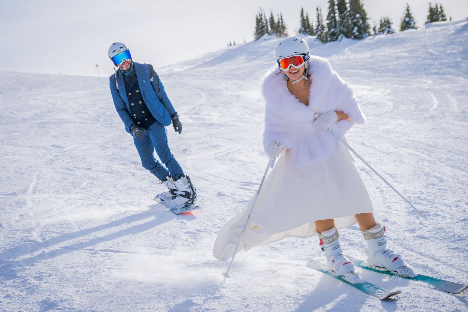Two skiers in winter attire gliding down a snowy mountain slope on a bright sunny day.