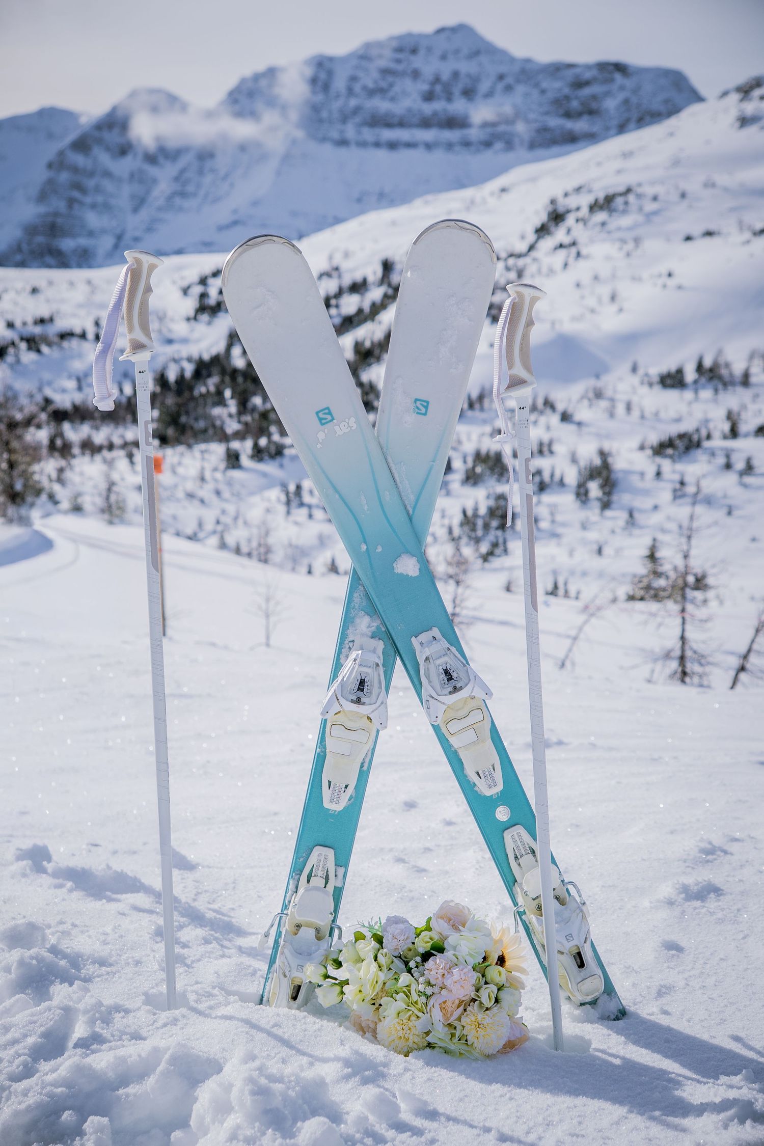 Turquoise skis stand crossed in snowy mountain landscape with white flowers at their base against dramatic alpine peaks.