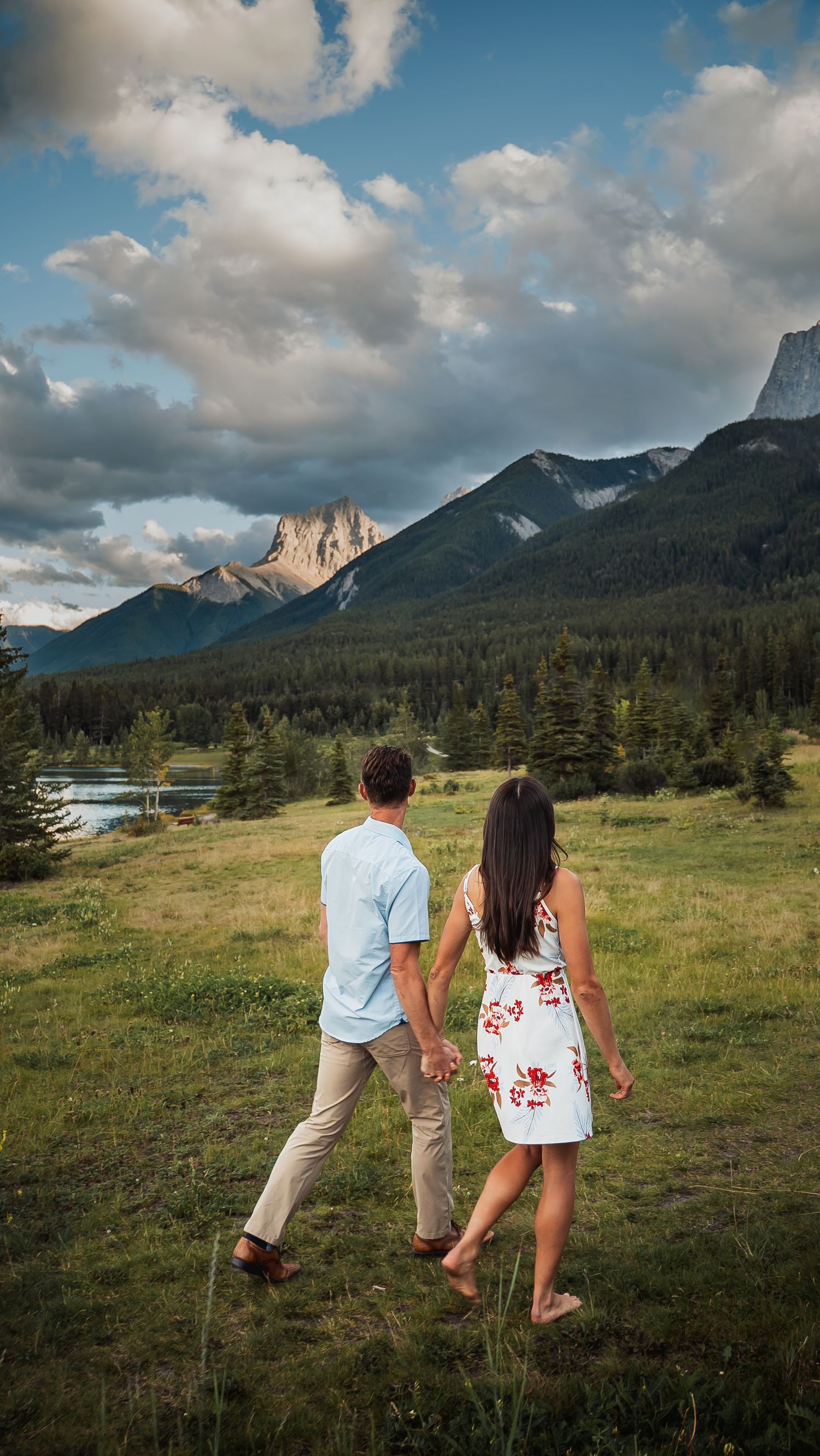 A couple holds hands while walking through a scenic mountain meadow with evergreen trees and peaks in the distance.