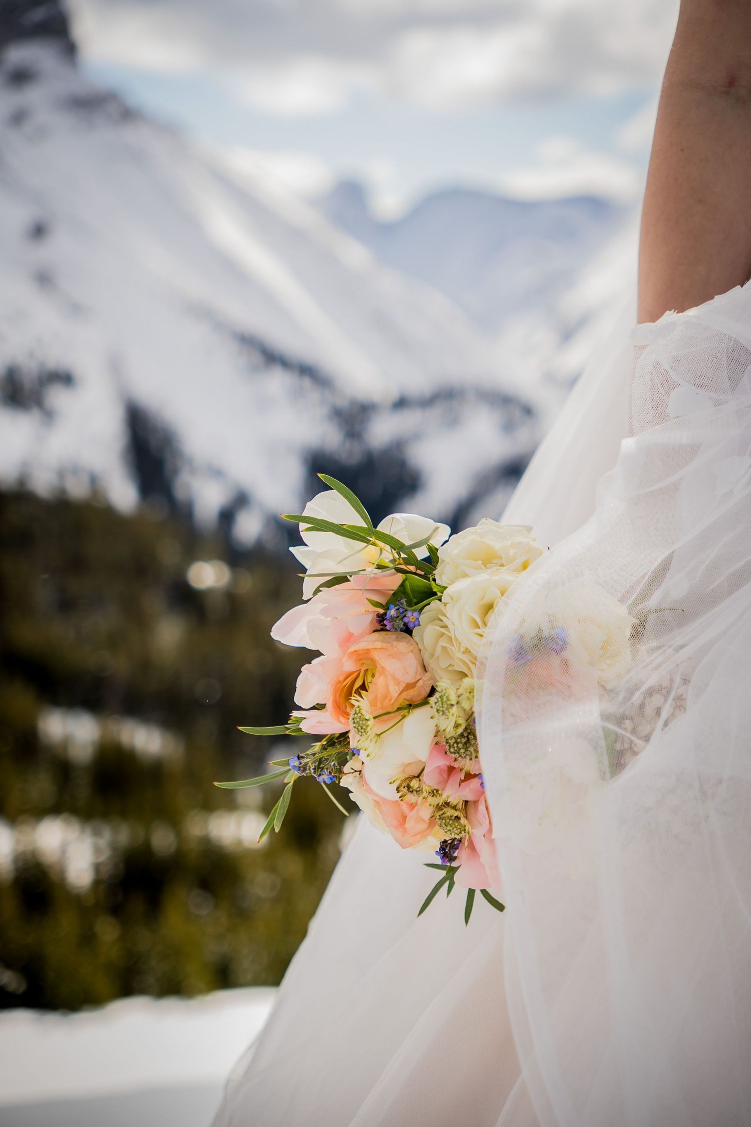 Bride holding a pastel pink and white floral bouquet with snowy mountain peaks in the background at a winter wedding.