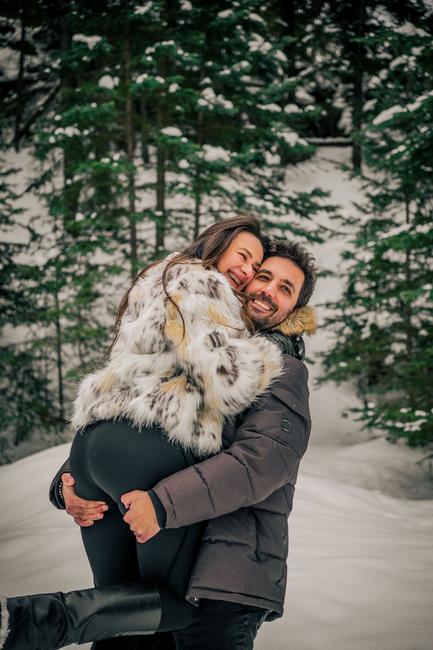 Couple embracing in snowy forest wearing winter coats.