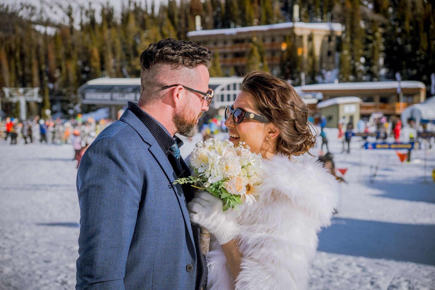 A couple shares a romantic moment during their winter wedding at a snowy ski resort with evergreen trees in the background.