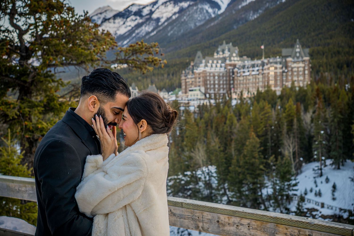 Couple shares intimate moment with majestic Canadian Rockies and Banff Springs Hotel visible in winter setting.