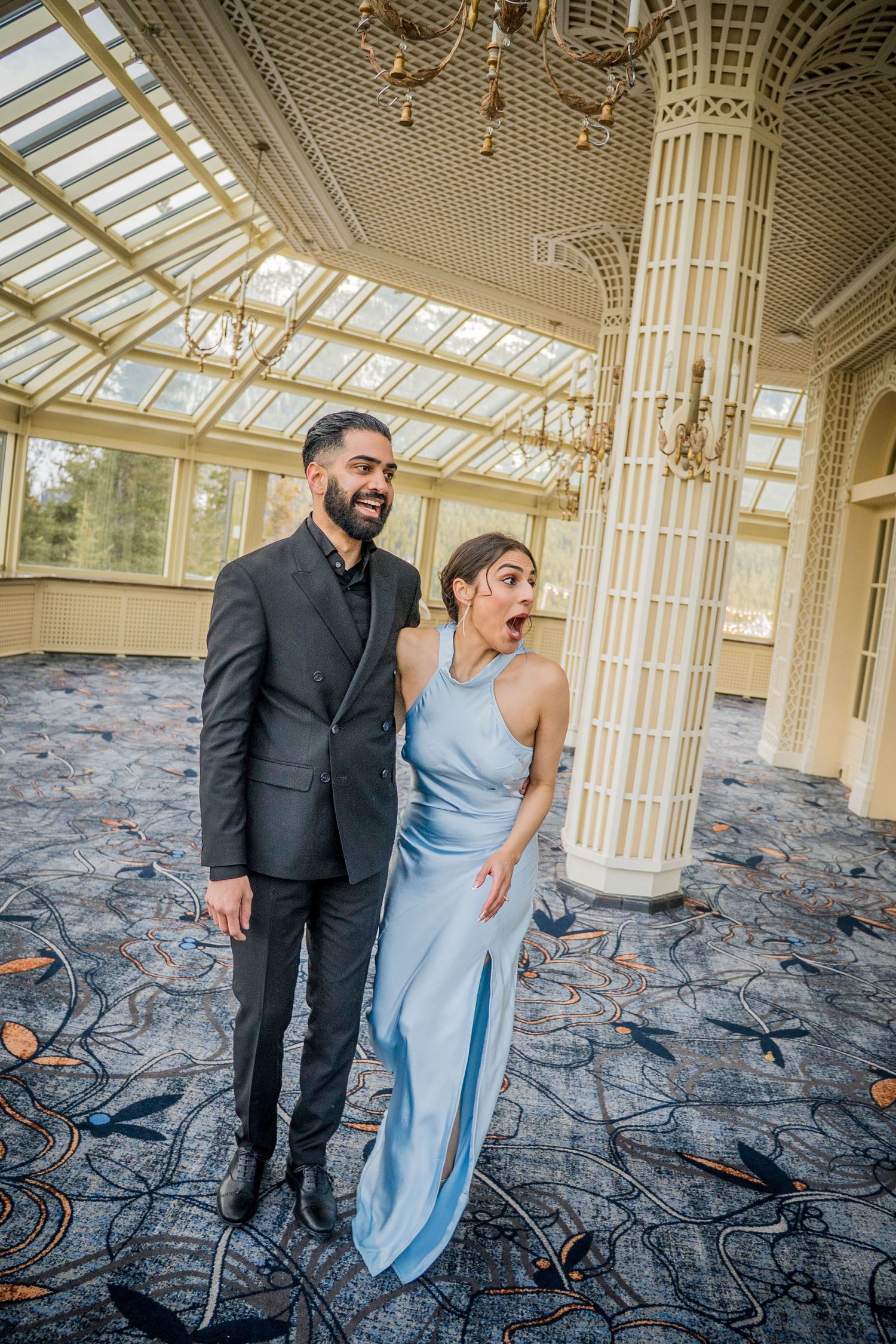 A couple in formal attire share candid moments and laughter in an elegant room with ornate columns and skylights.