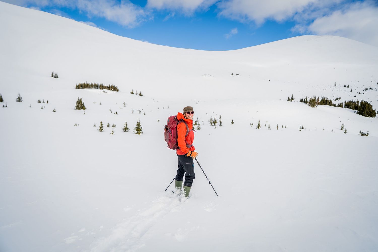 A skier in an orange jacket with red backpack and poles traverses a snowy mountain slope under blue skies.
