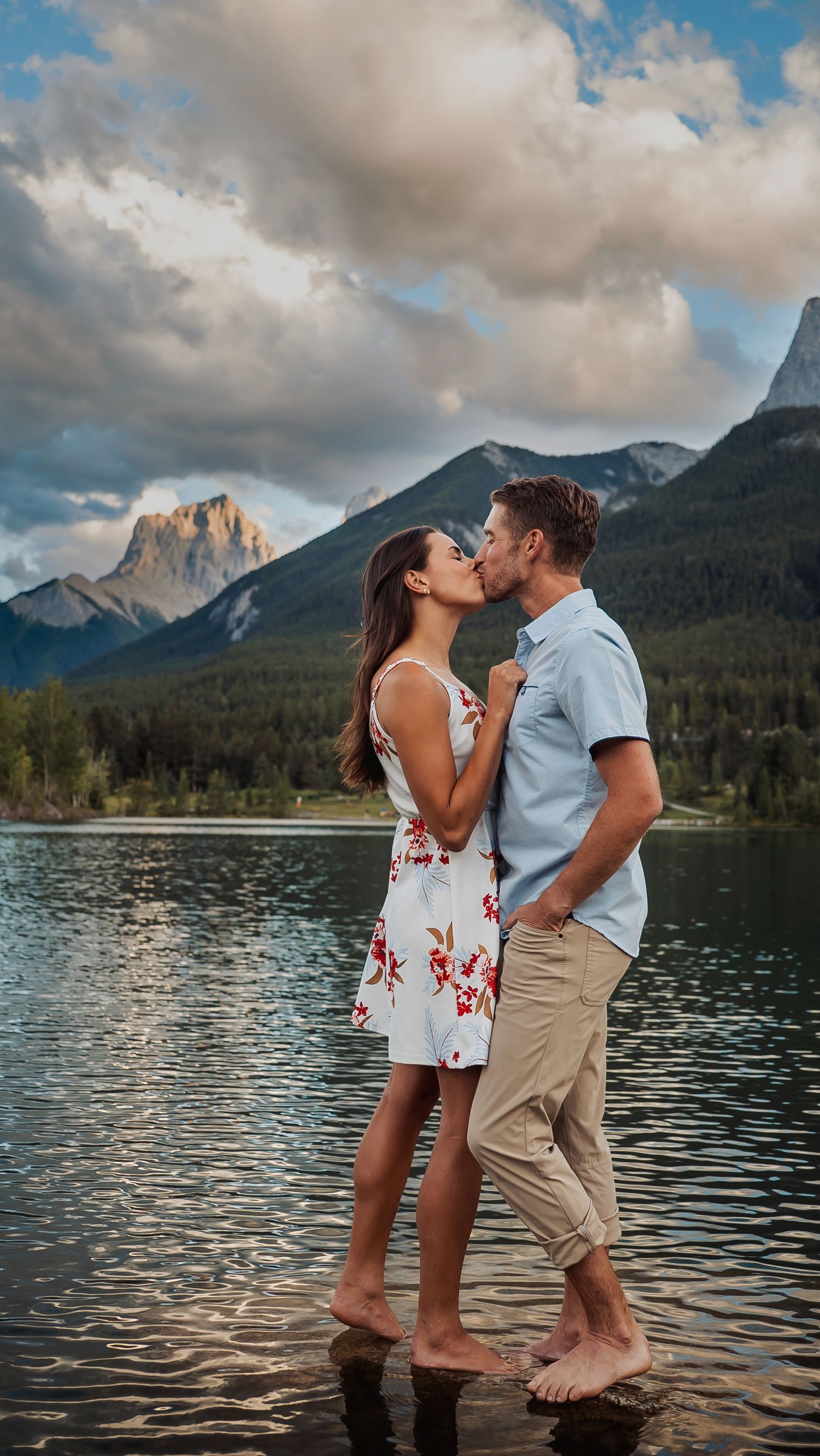 A romantic couple shares a kiss by a mountain lake at sunset with dramatic cloudy skies and peaks in the background.