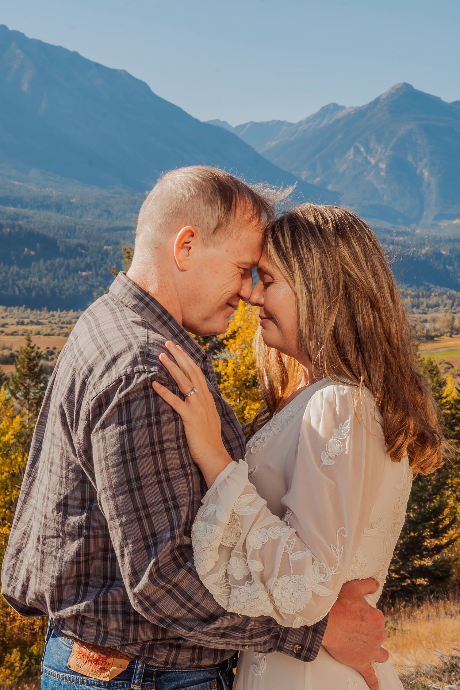A romantic couple embraces against a backdrop of majestic mountain peaks and autumn foliage in golden sunlight.
