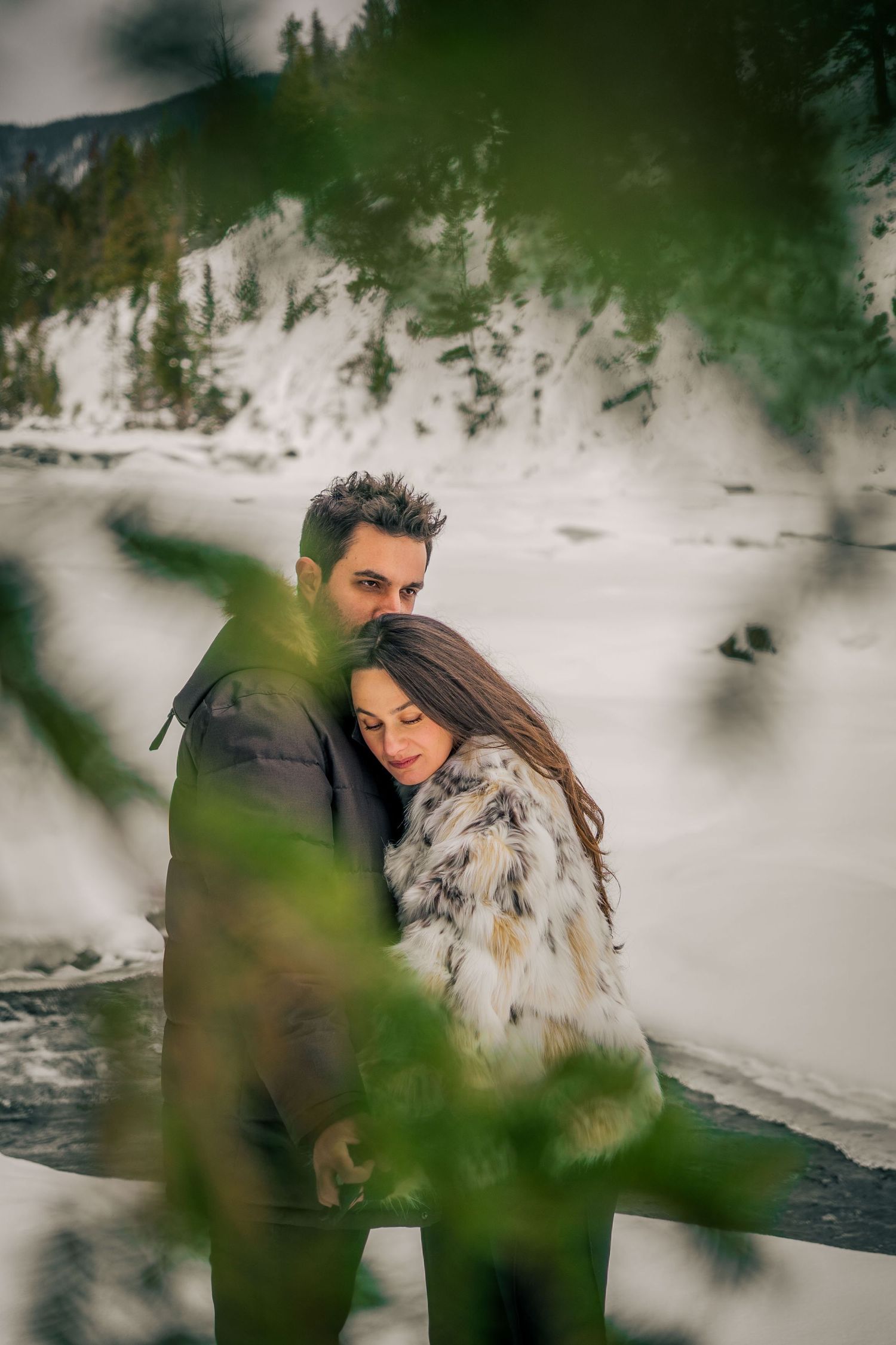 Winter mountain scene with couple embracing viewed through pine branches.