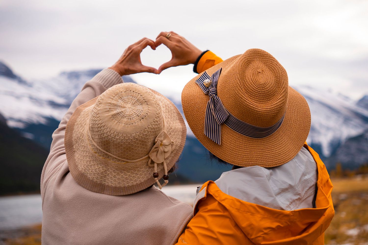 Two people wearing sun hats making heart shapes with hands against mountain backdrop.