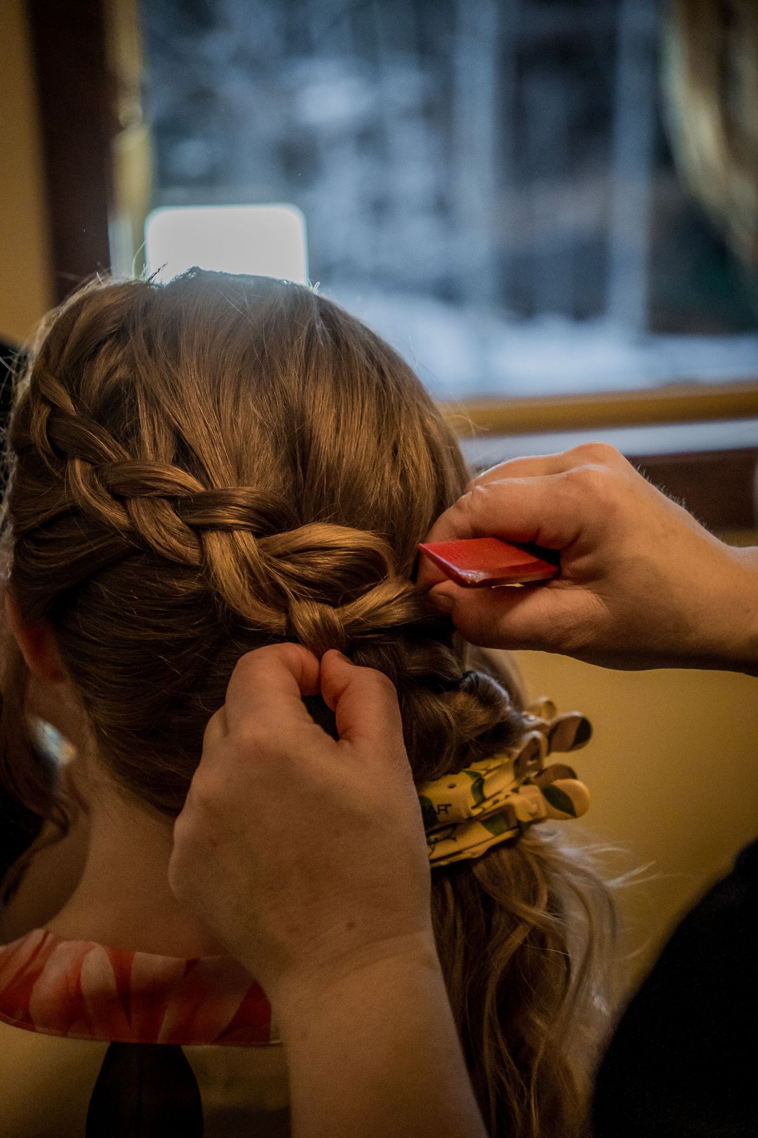 Hairstylist braiding a woman's hair into an elegant updo, securing with a red clip in a warm indoor setting.