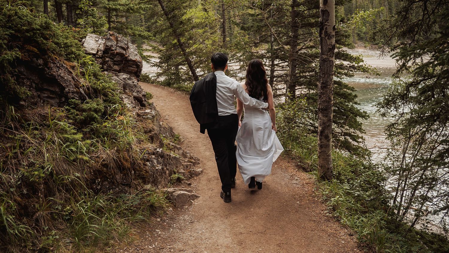 People walking along a scenic forest trail near a tranquil lake with tall evergreen trees surrounding the path.