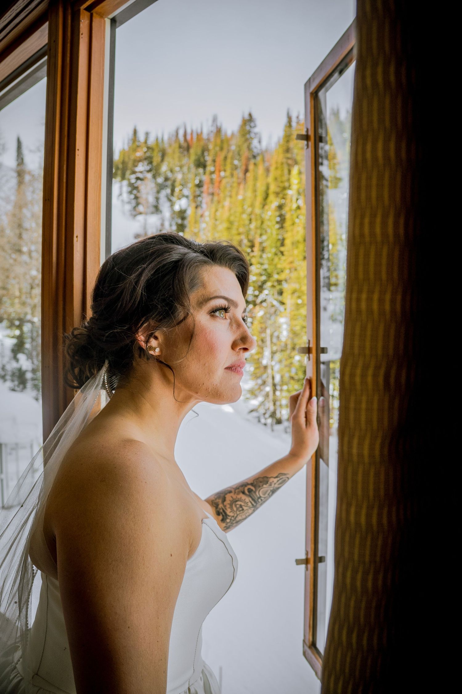 Bride in white dress gazes thoughtfully out window at snowy winter landscape with evergreen trees.
