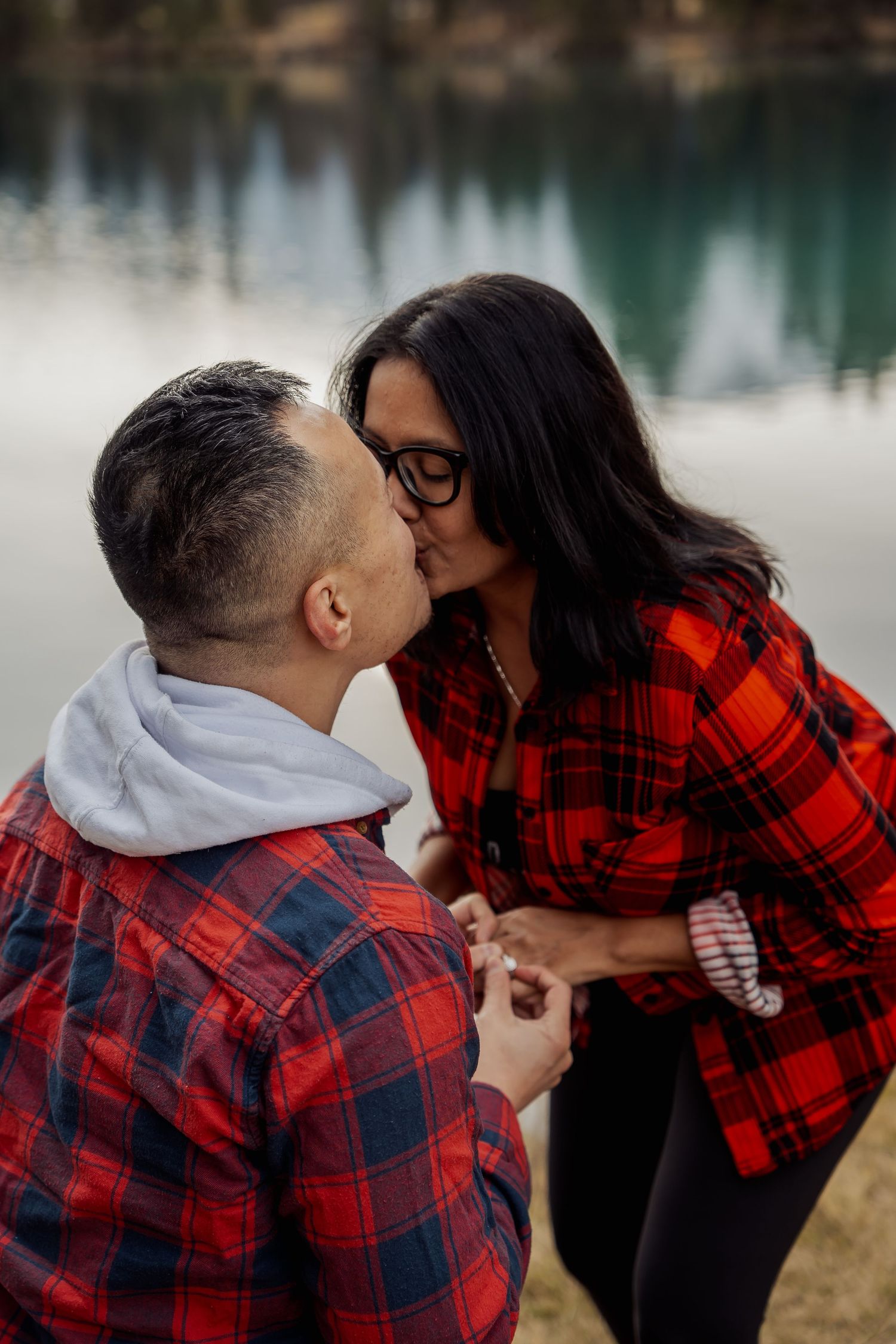 A couple in red plaid clothing embraces near a peaceful lake.