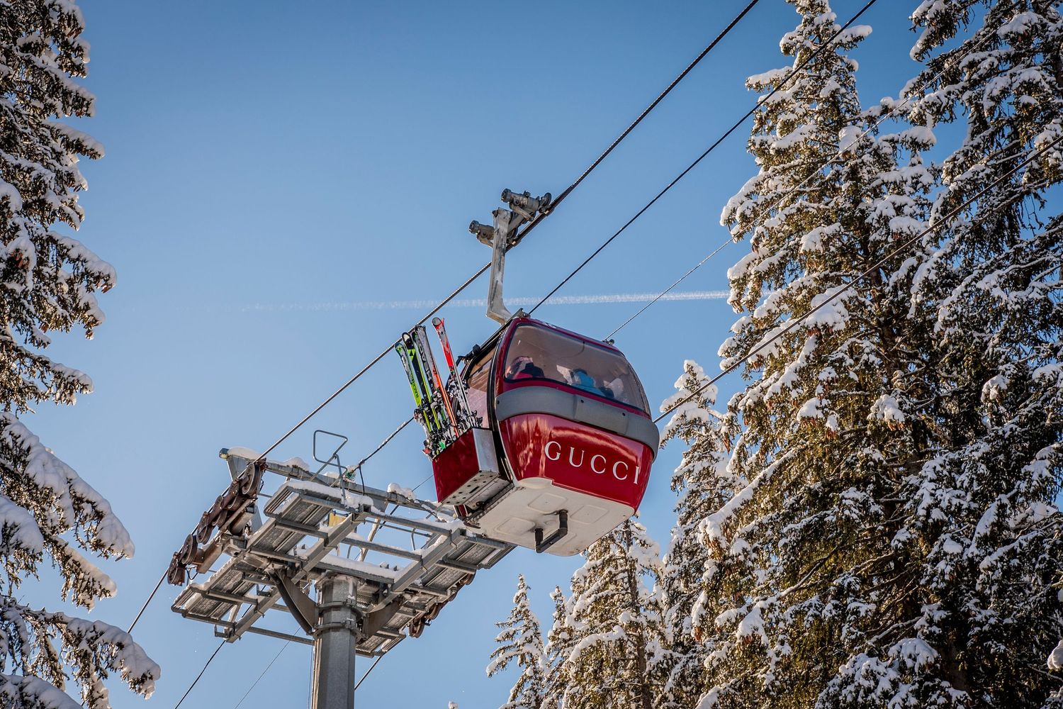 A red ski gondola lift moves through snow-covered pine trees against a bright blue winter sky.
