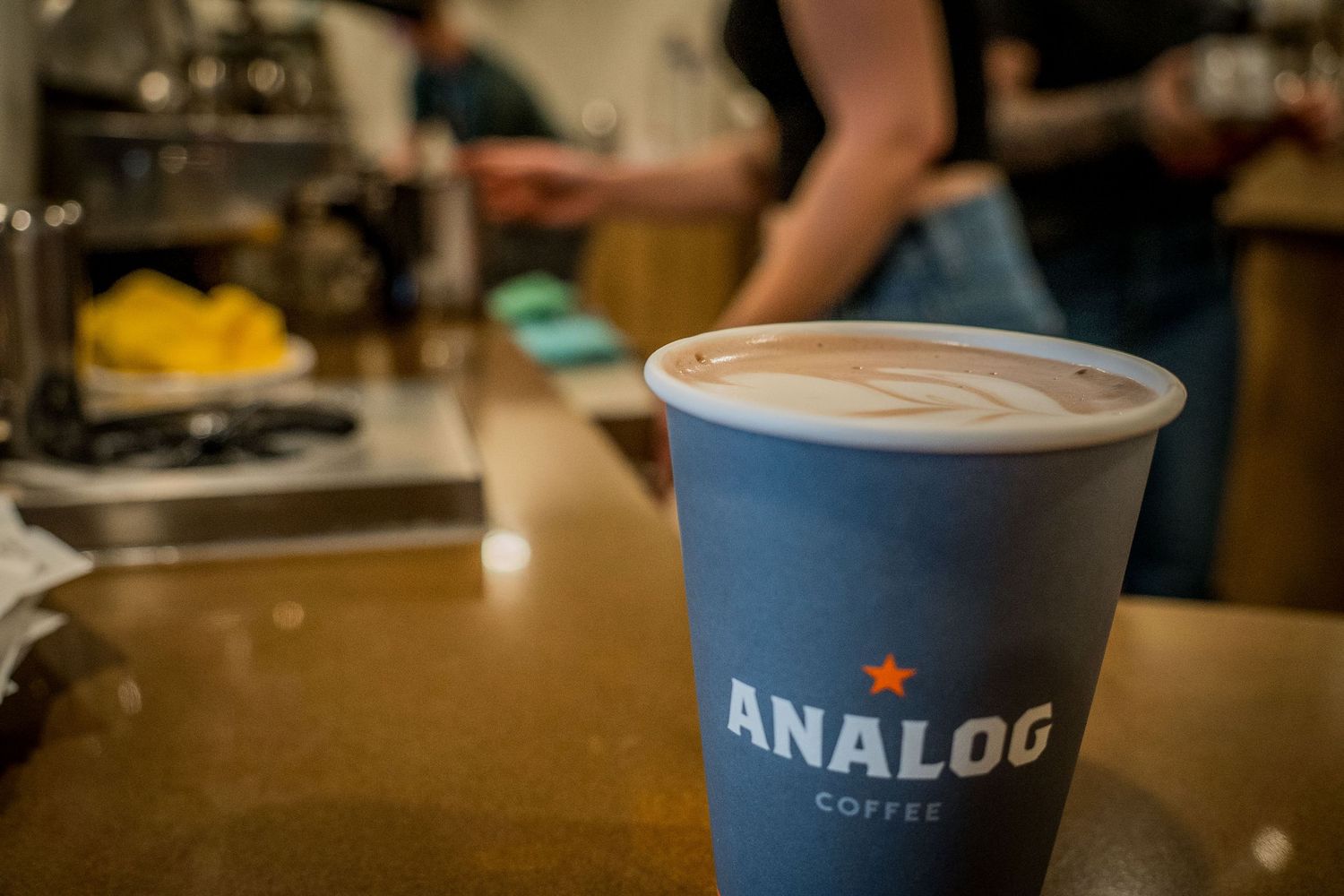 A coffee cup from Analog Coffee sits on a wooden table in a dimly lit cafe setting.