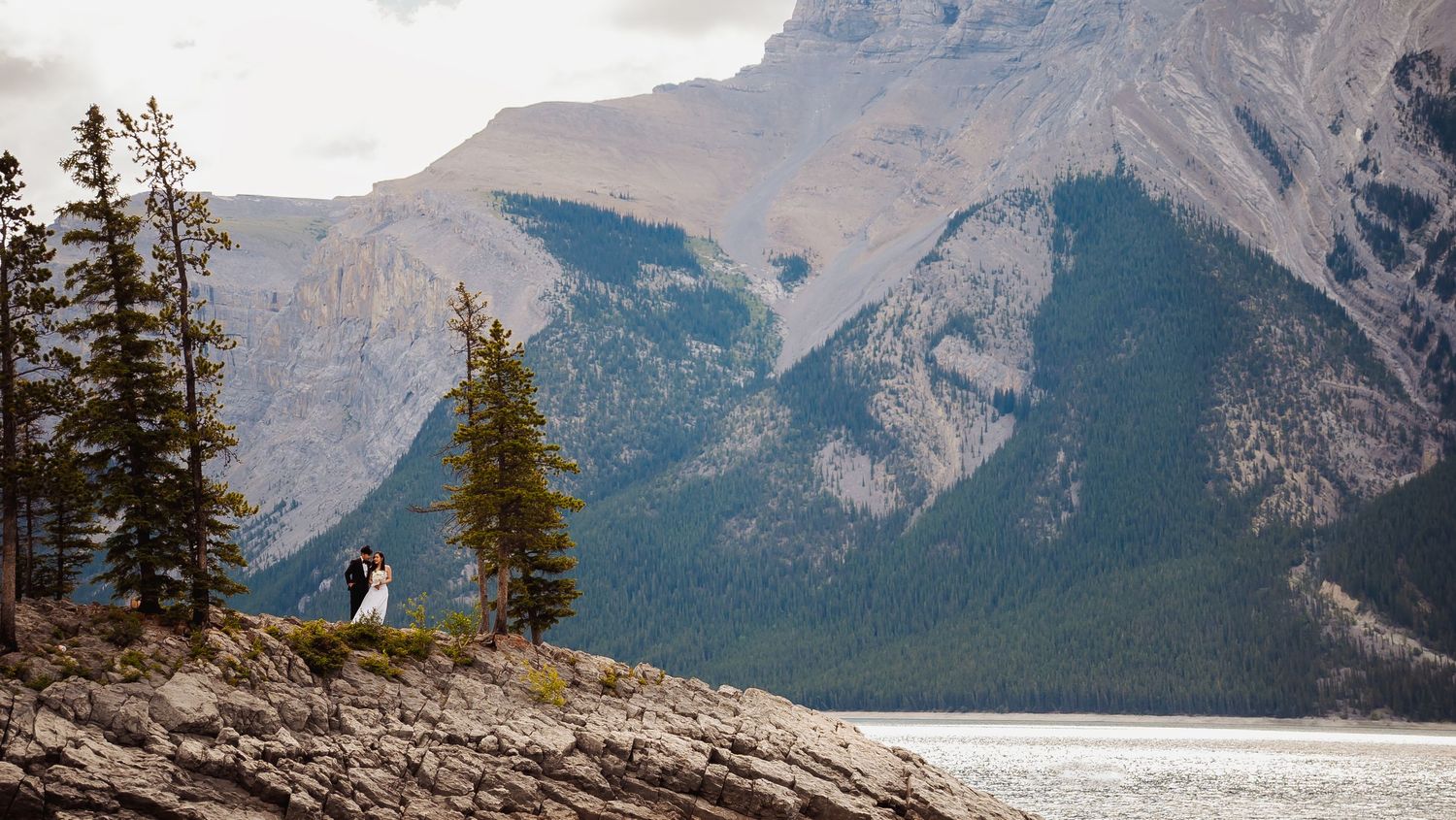 A couple stands on a rocky mountain ledge overlooking Lake Minnewanka in Banff National Park, surrounded by majestic peaks.