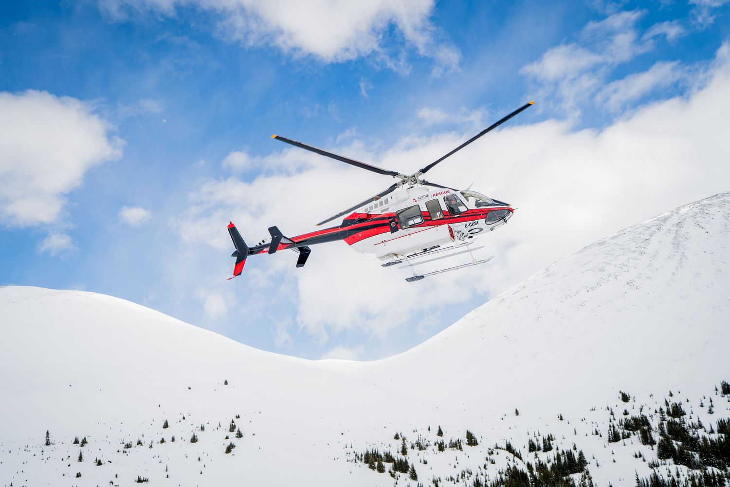 Red and white helicopter flying over snow-covered mountain peaks against a blue sky with clouds.