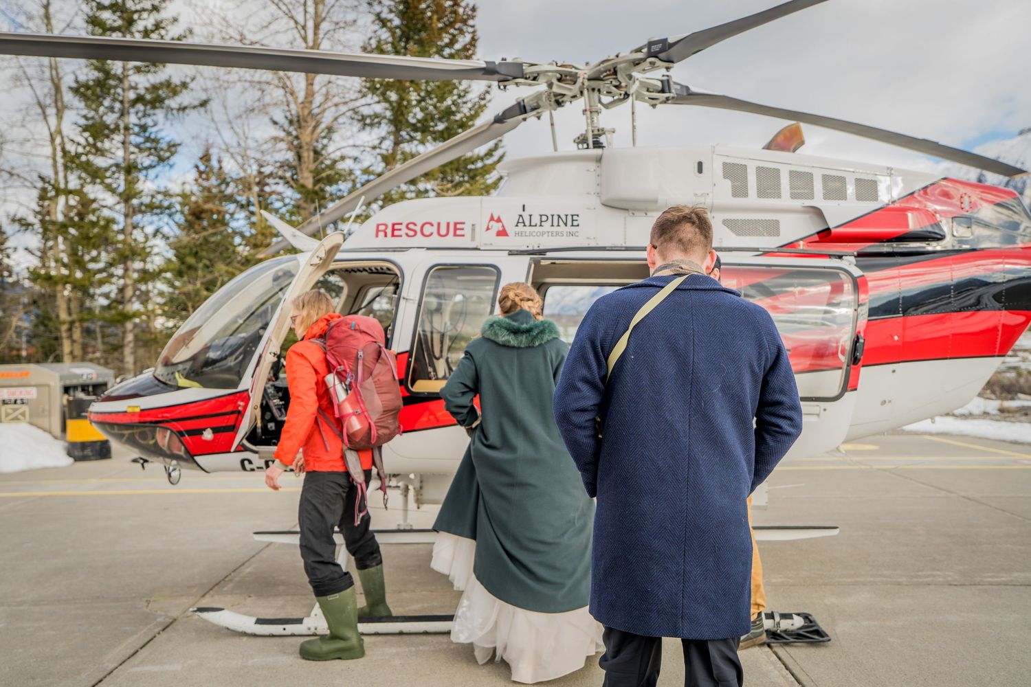 Group of people boarding an Alpine Rescue helicopter on a snowy helipad surrounded by winter trees.