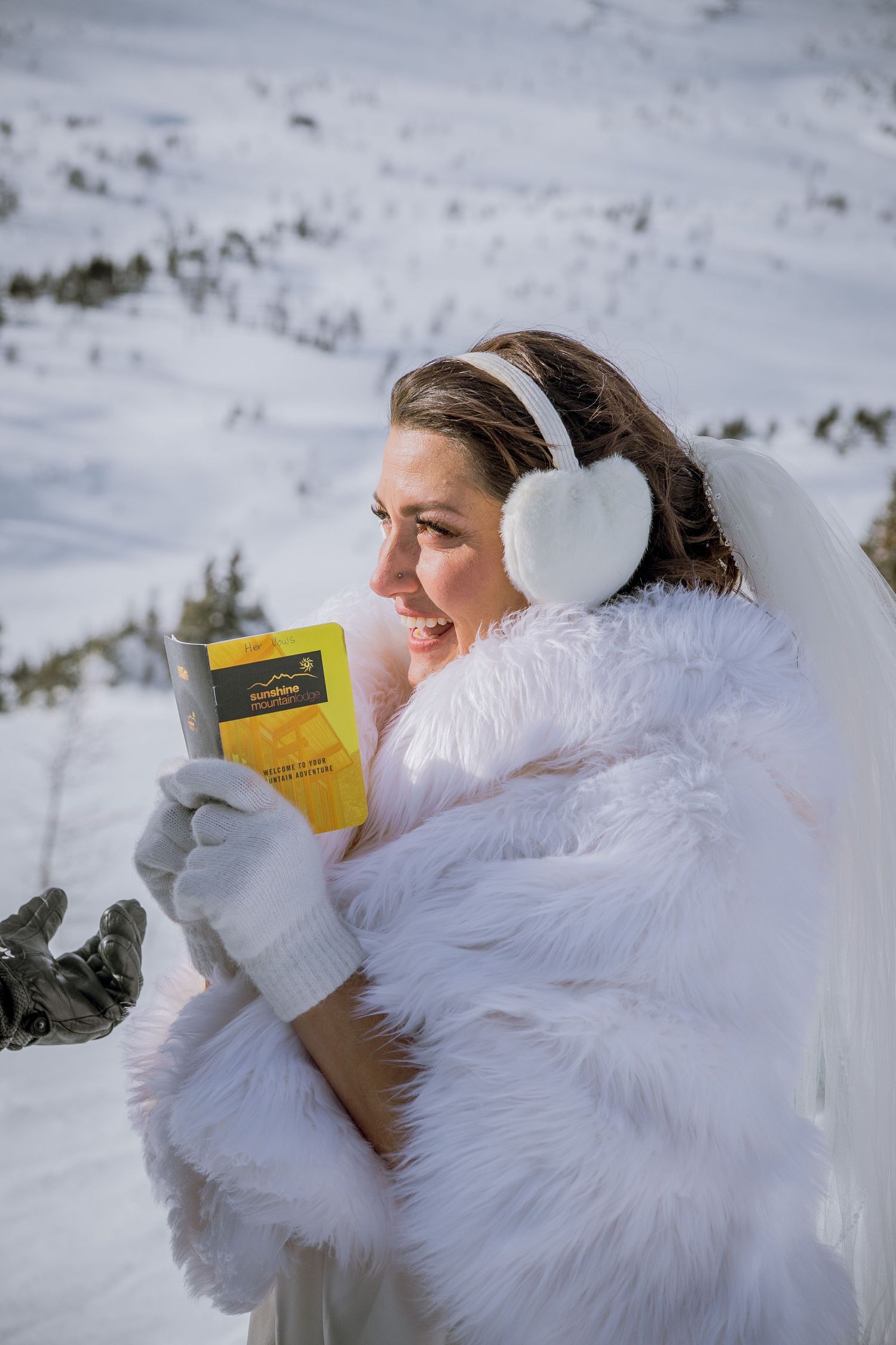 Person in fluffy white winter coat holds yellow book against snowy mountain backdrop.