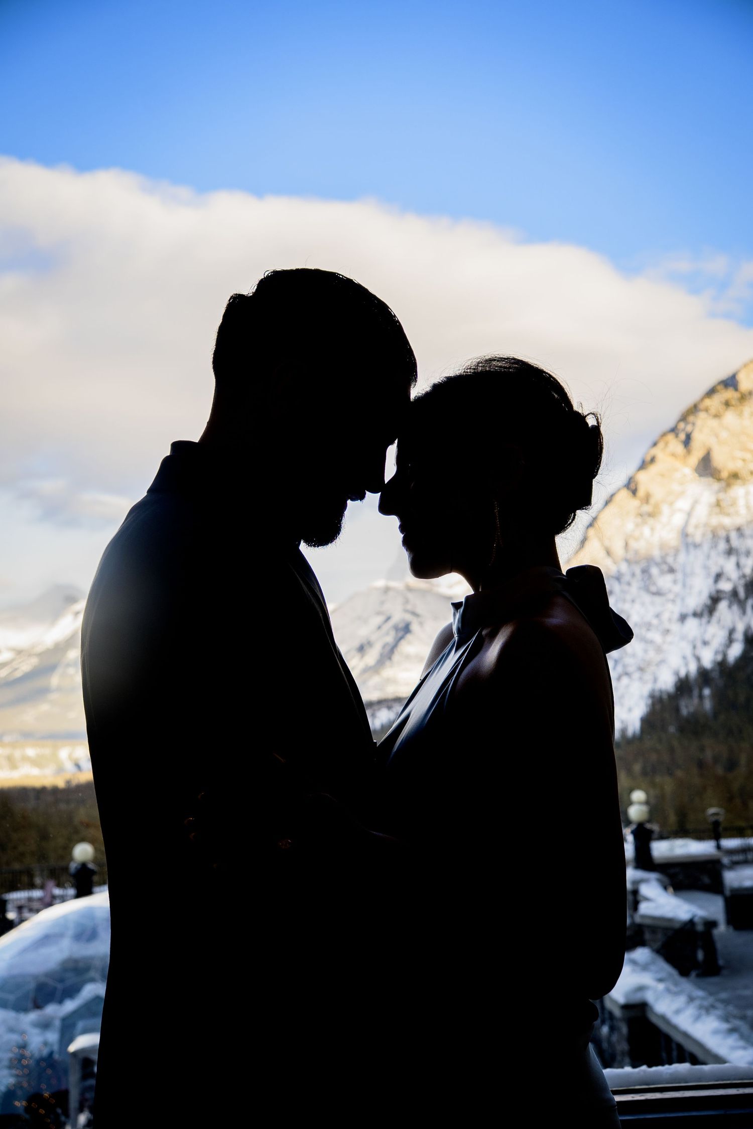 A silhouetted couple shares an intimate moment against a snowy mountain backdrop with blue sky and clouds.