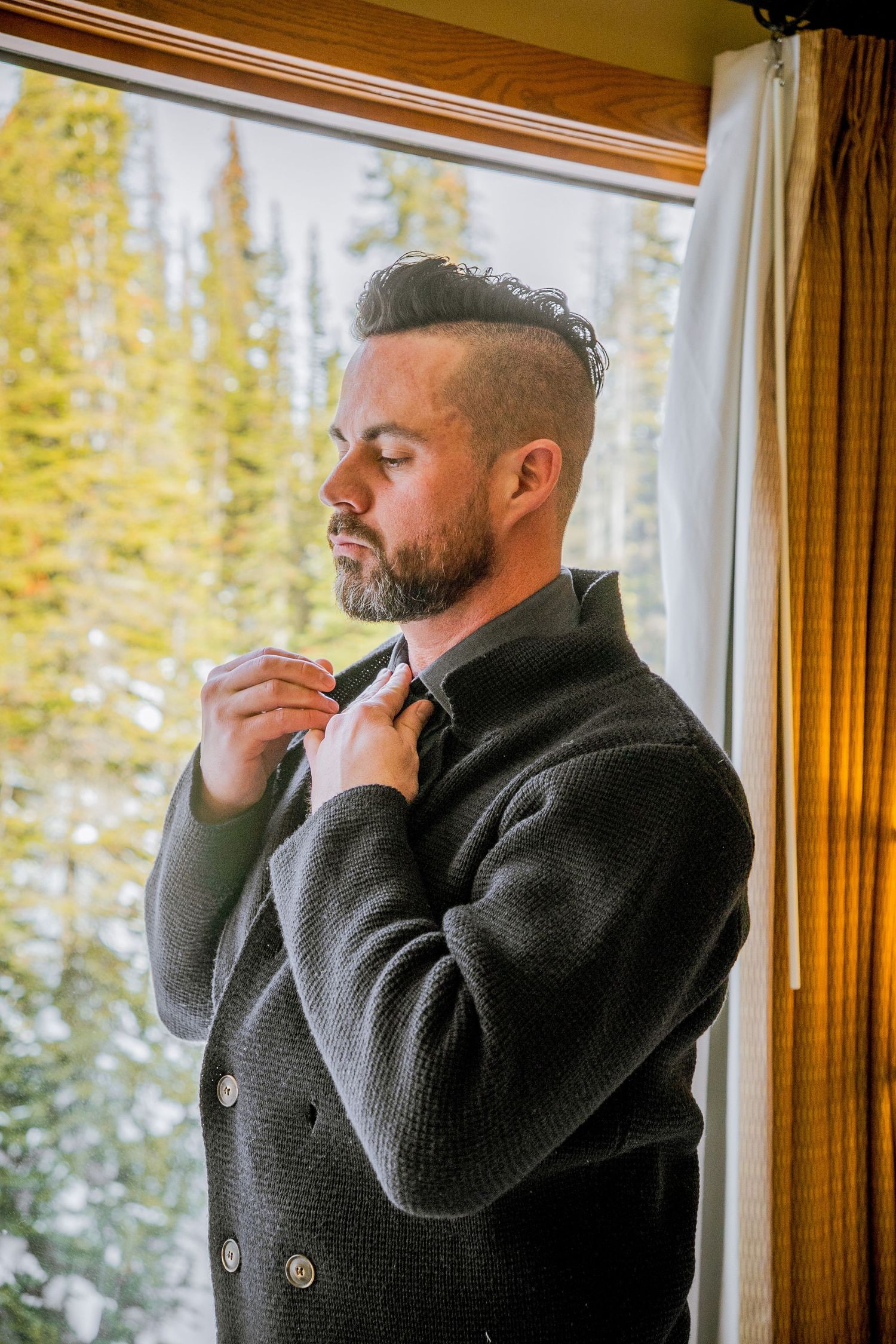 Man in black turtleneck sweater adjusts collar while standing by window with autumn trees visible outside.