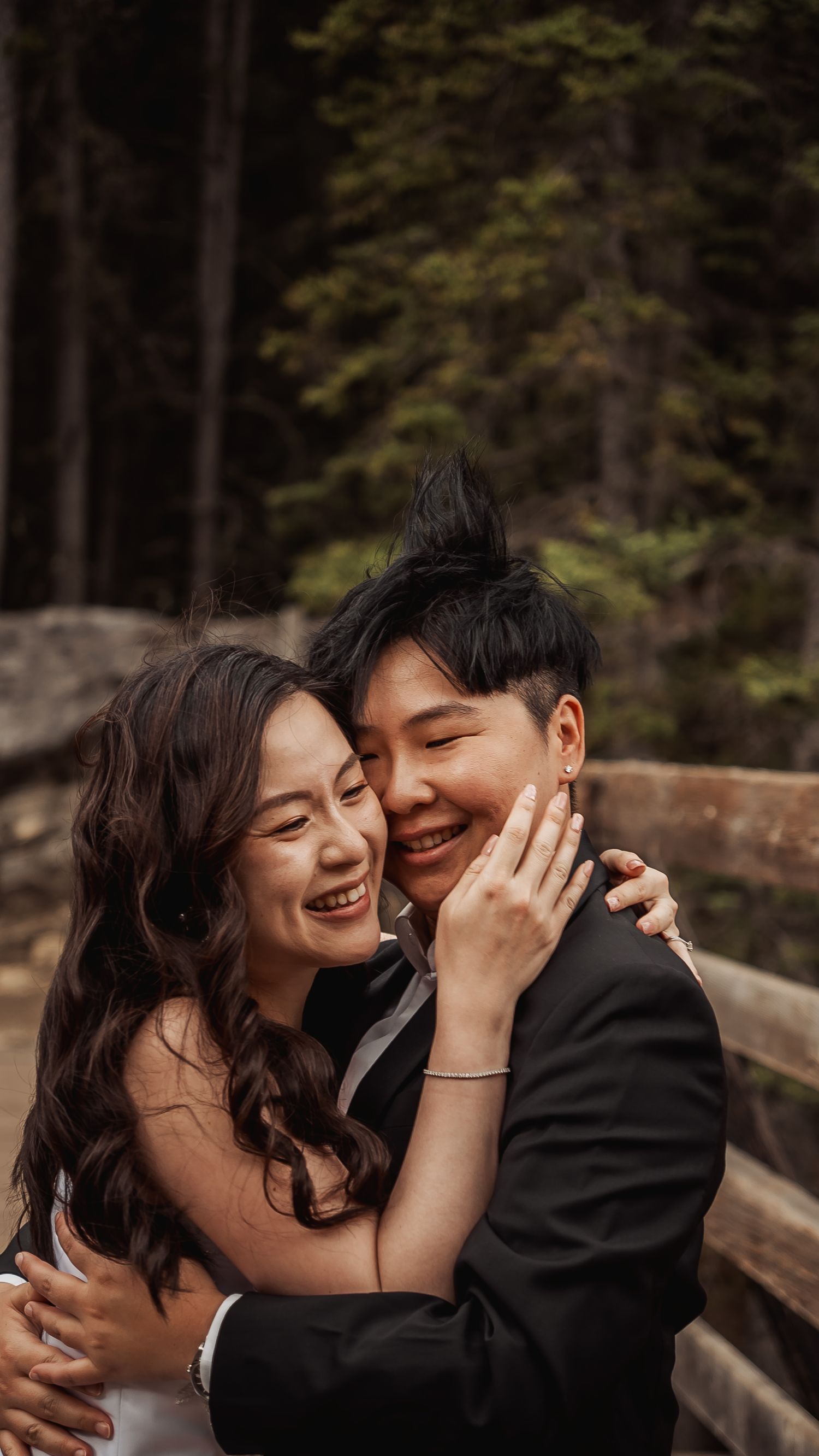 A joyful couple embraces and laughs together on a wooden bridge surrounded by forest scenery.