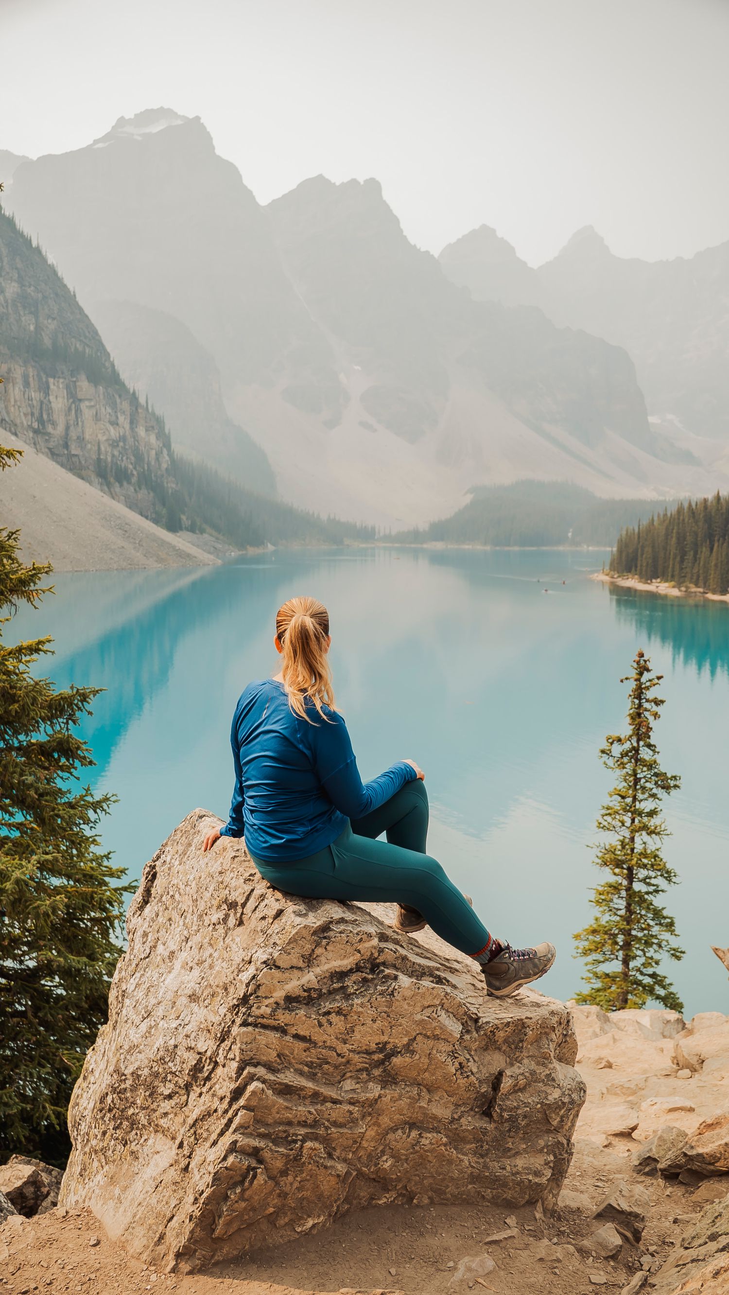 Person in blue sitting on rock overlooking turquoise mountain lake with peaks in background.