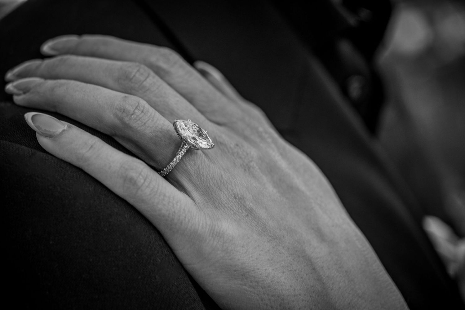 Close-up black and white photograph of a diamond engagement ring on a hand against a dark background.