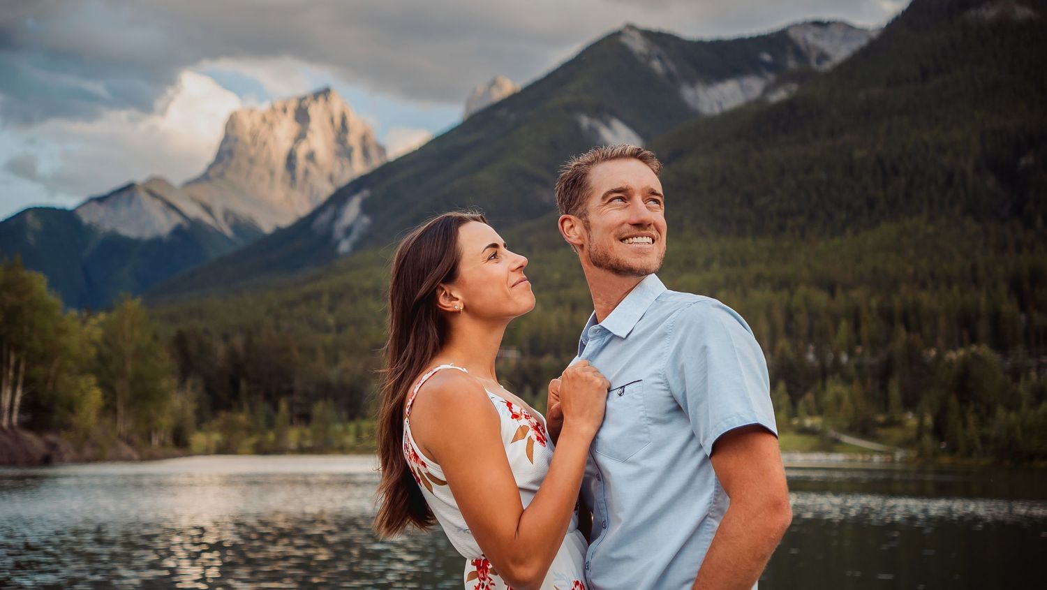 A couple poses together in front of scenic mountain landscapes and lakes during an outdoor engagement photoshoot.