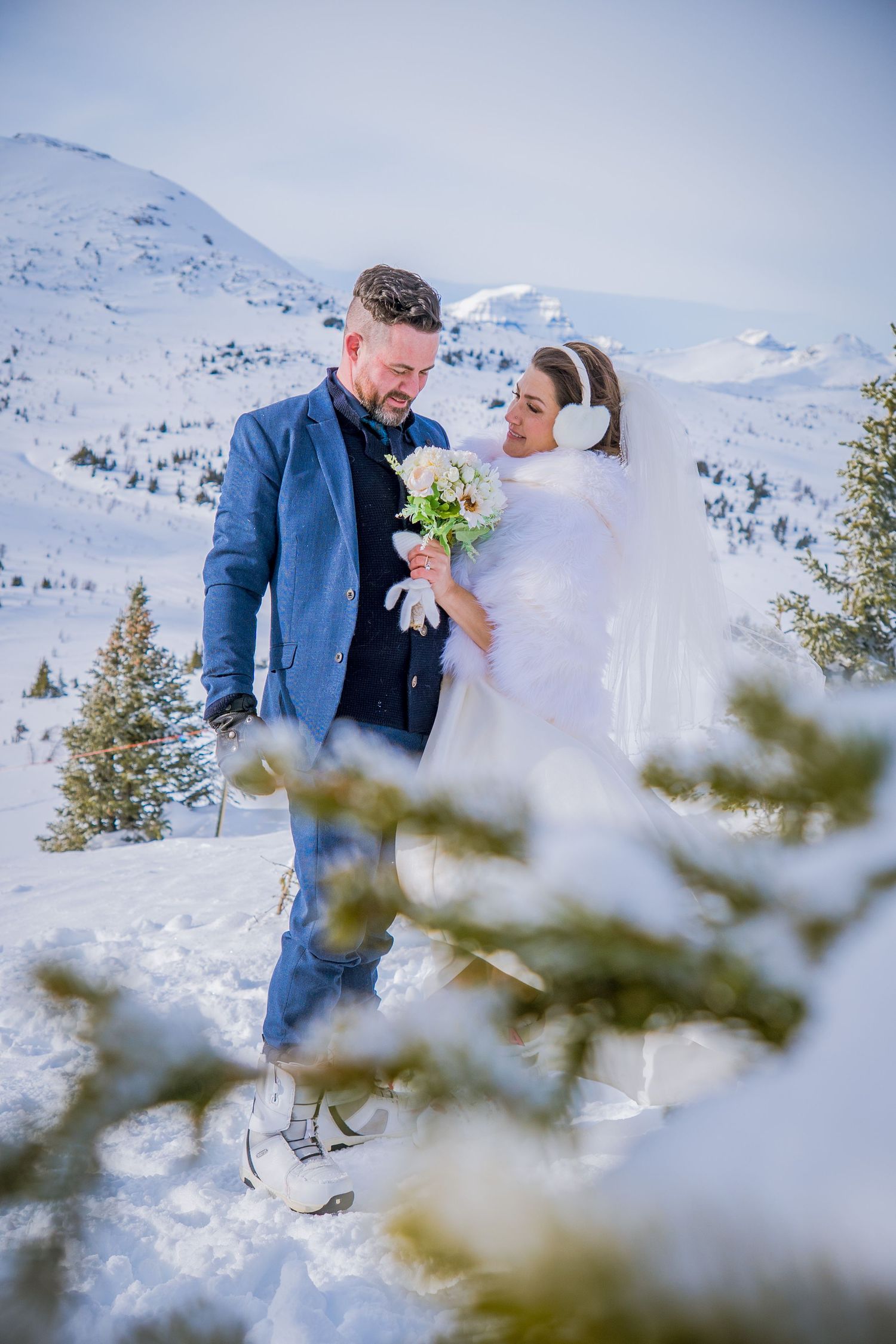 A couple shares an intimate embrace surrounded by snow-covered evergreens in a mountainous winter landscape.