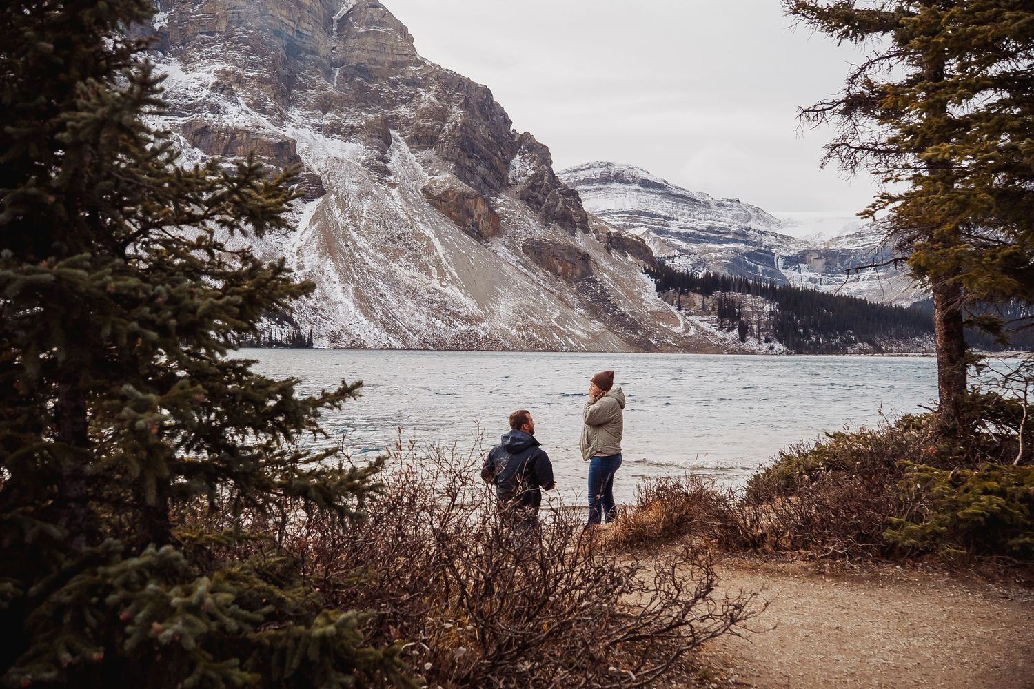 Two people stand by a frozen mountain lake surrounded by snow-dusted peaks and evergreens.