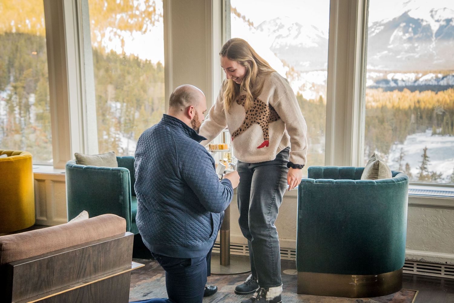 Two people share a tender moment in a modern living room with large windows overlooking autumn mountain scenery.
