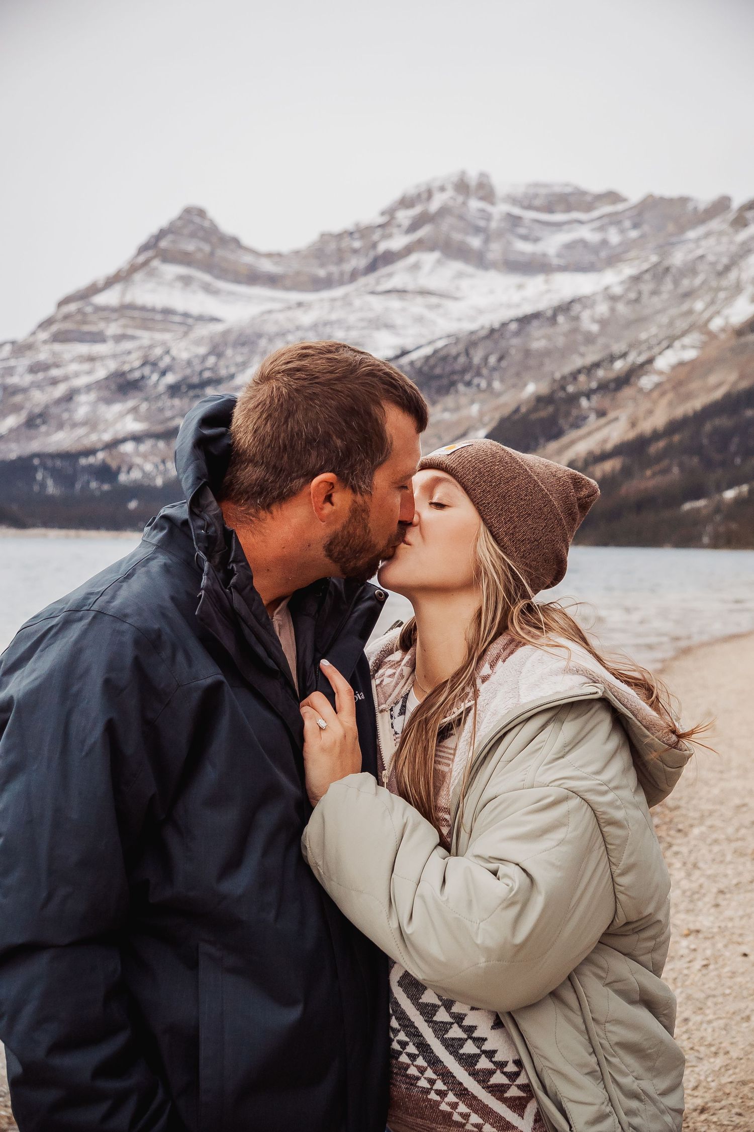A couple in winter clothing shares intimate kisses against a backdrop of snowy mountain peaks.