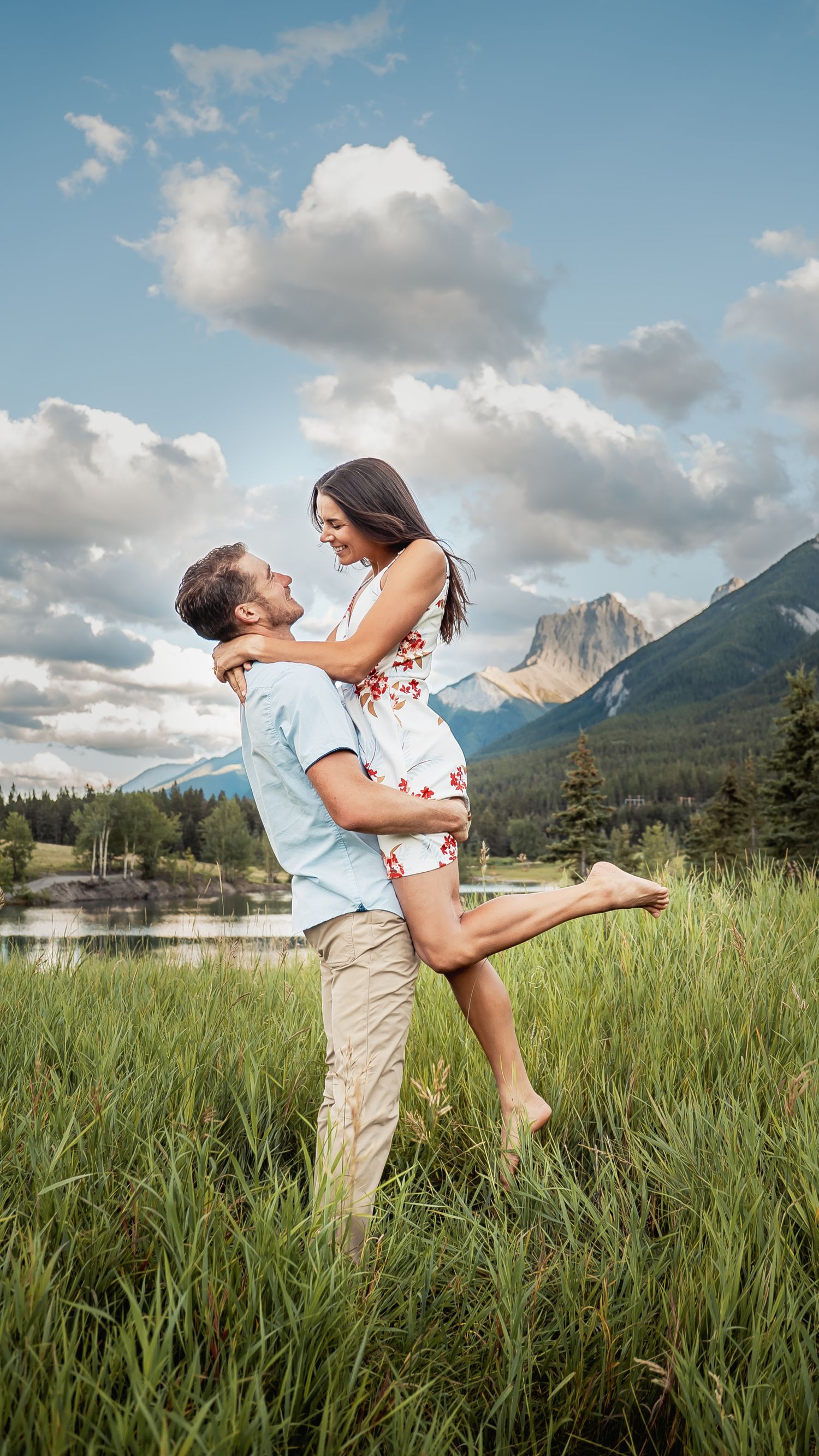 Romantic couple embraces in a grassy meadow with majestic mountain peaks and blue skies in the background.