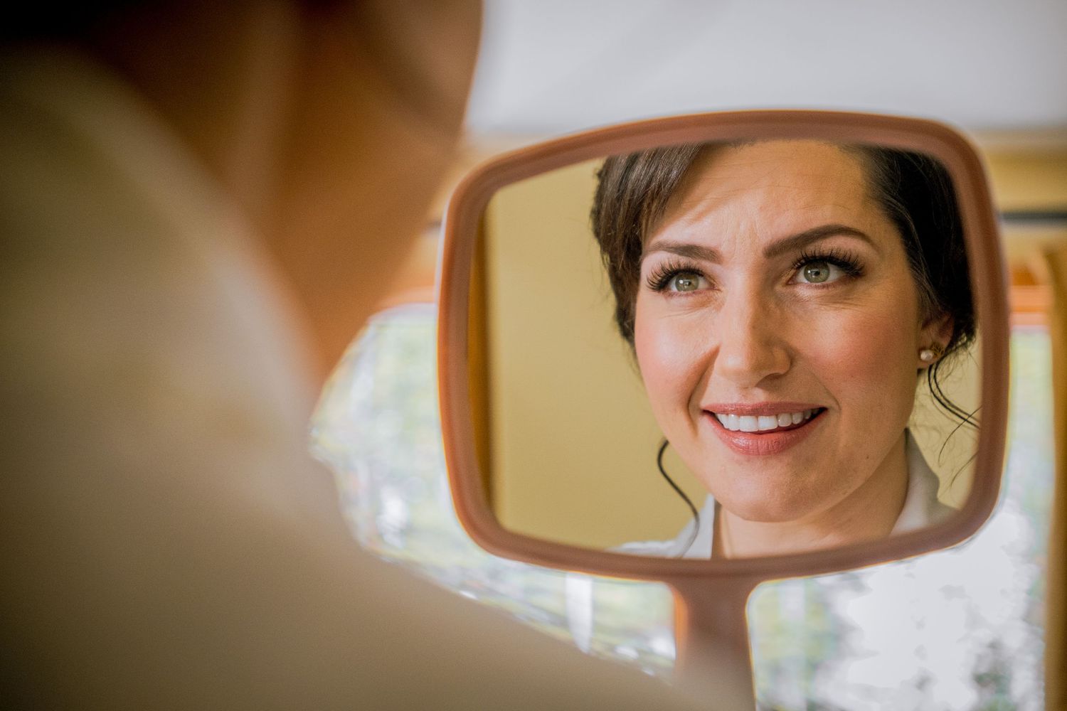 A bride gets her hair and makeup done while looking in a mirror during wedding preparations.