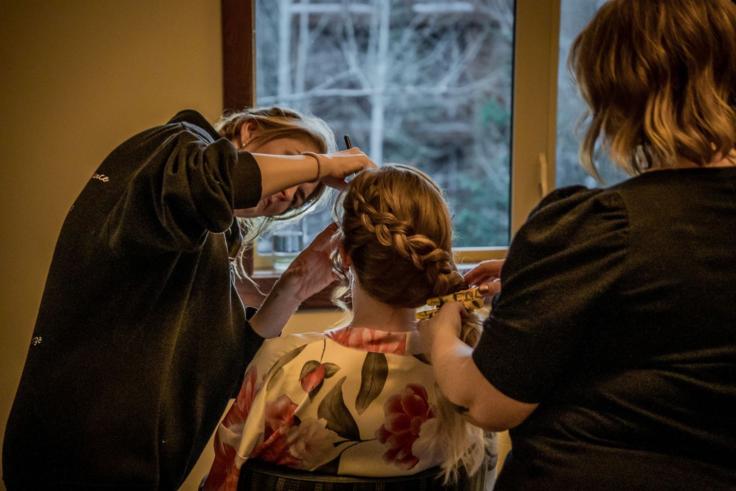 Two stylists carefully styling a womans elegant updo hairstyle near a window in a warmly lit room.