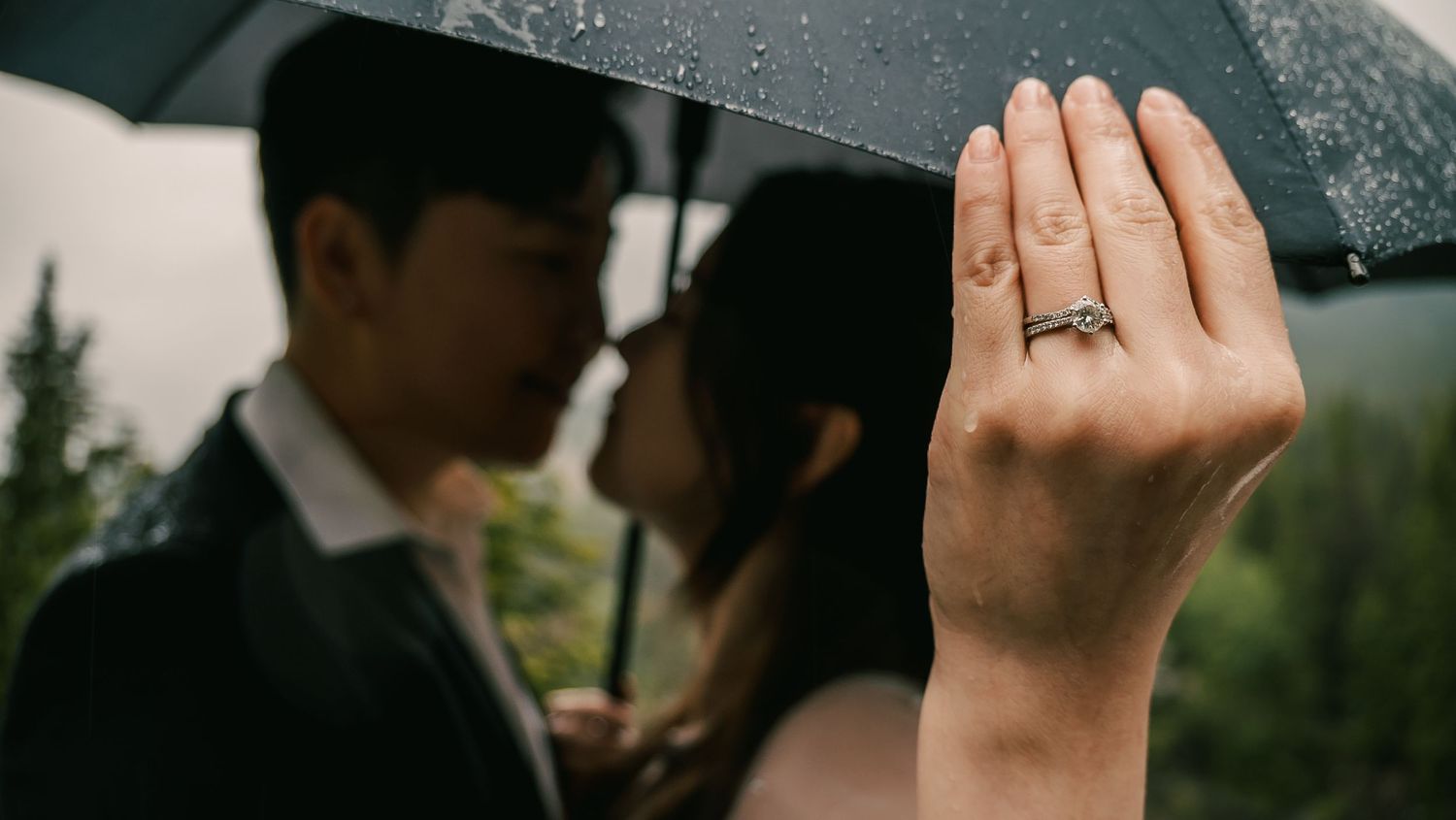 Close-up of an engagement ring on a hand with a couple holding an umbrella in the rainy background.
