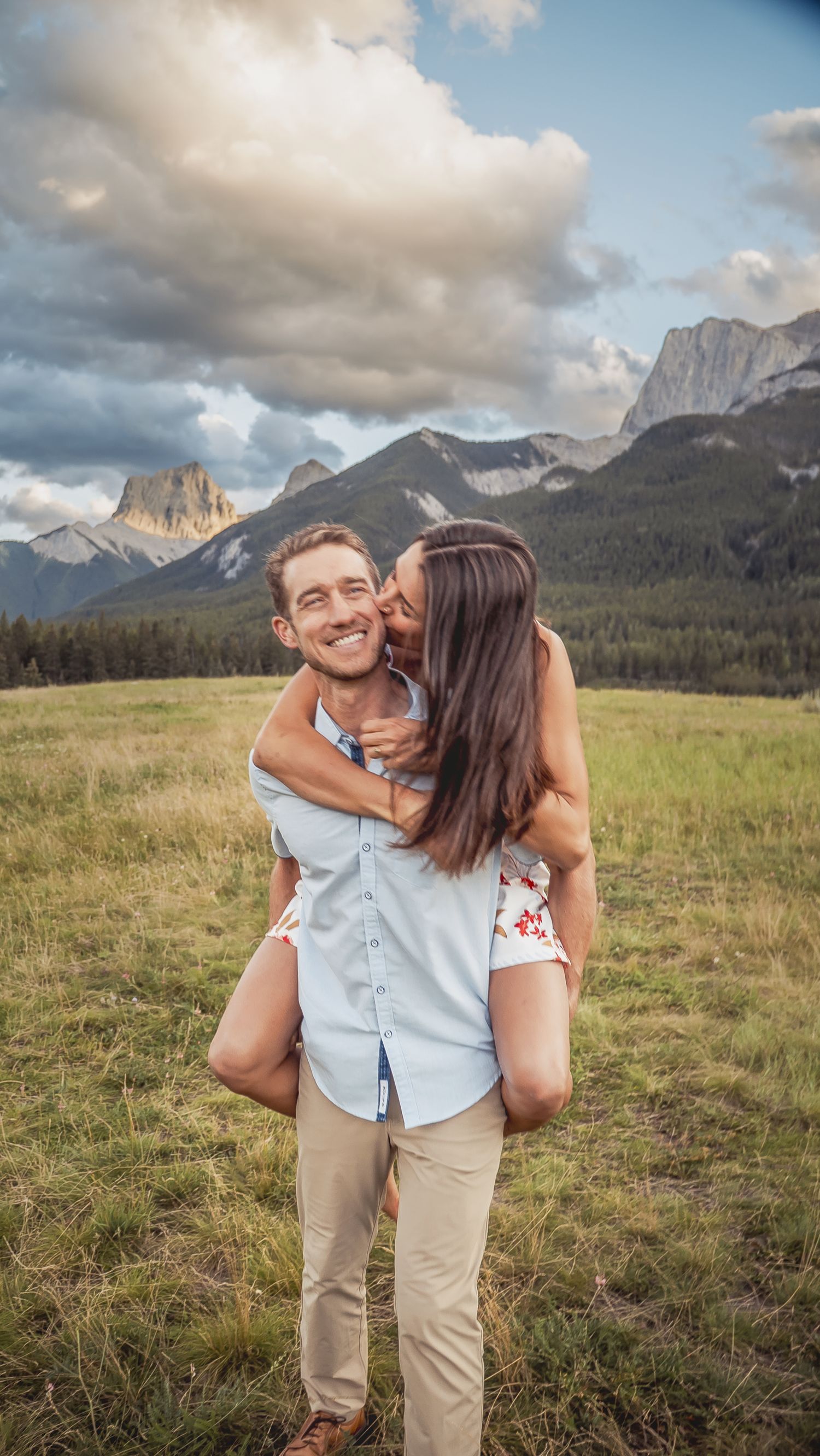 A couple shares a playful moment in a mountain meadow with dramatic rocky peaks and cloudy skies in the background.