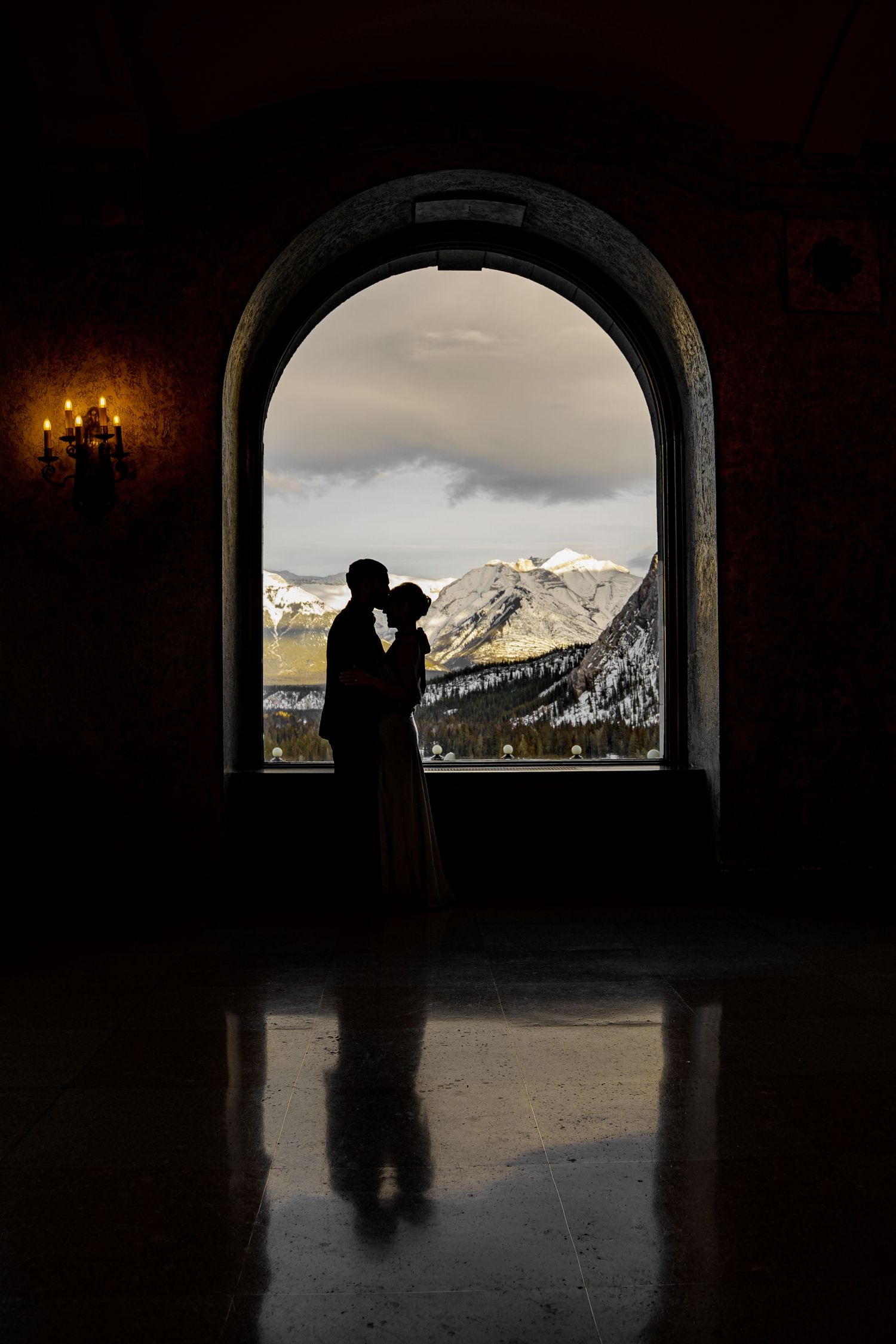 Silhouetted figures in an arched tunnel frame a dramatic mountain landscape with snow-capped peaks at sunset.