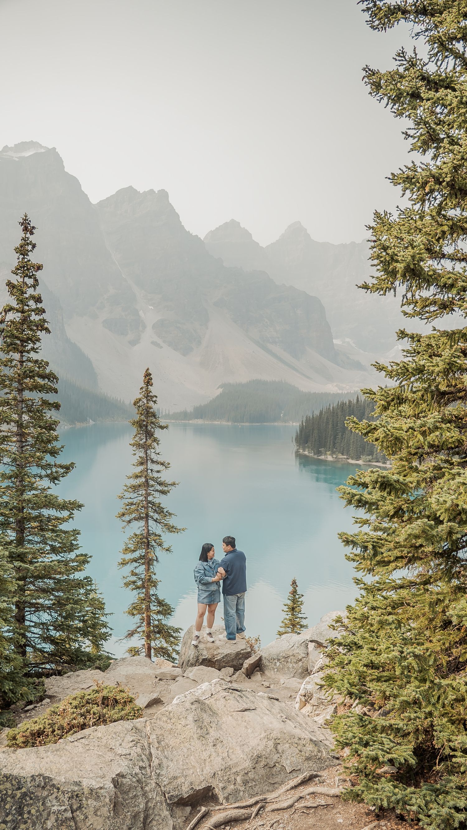 A couple stands on a rocky outcrop overlooking the stunning turquoise waters of Moraine Lake in Banff.