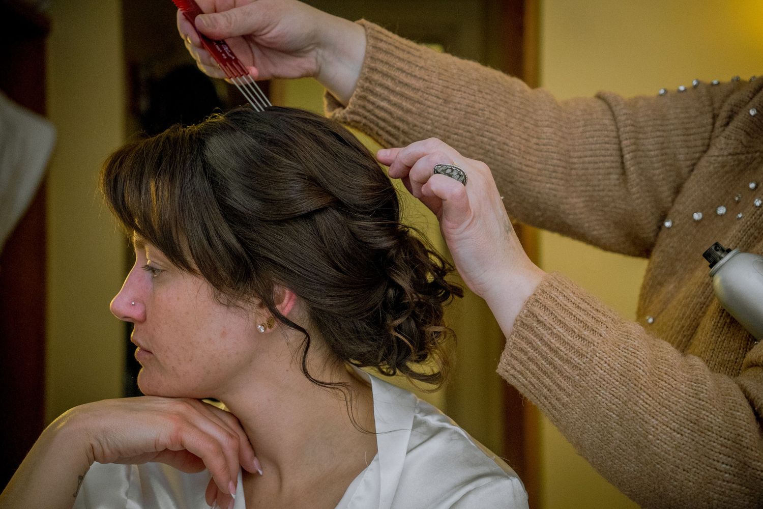 A makeup artist applies finishing touches to a bride's wedding day look in a warm-lit room.