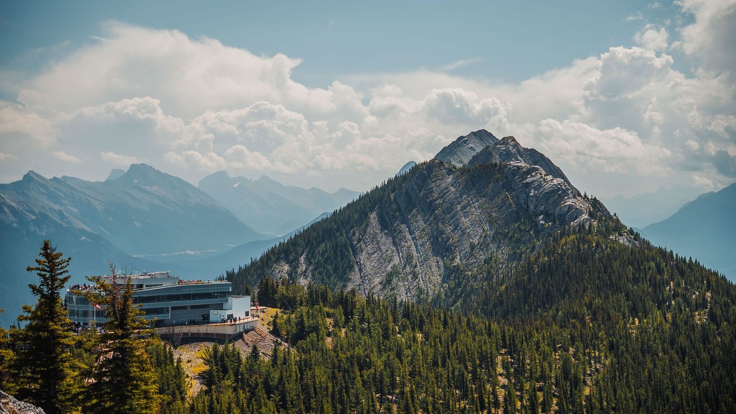 Panoramic view of snow-capped mountain peaks and evergreen forests under a bright blue sky with white clouds.