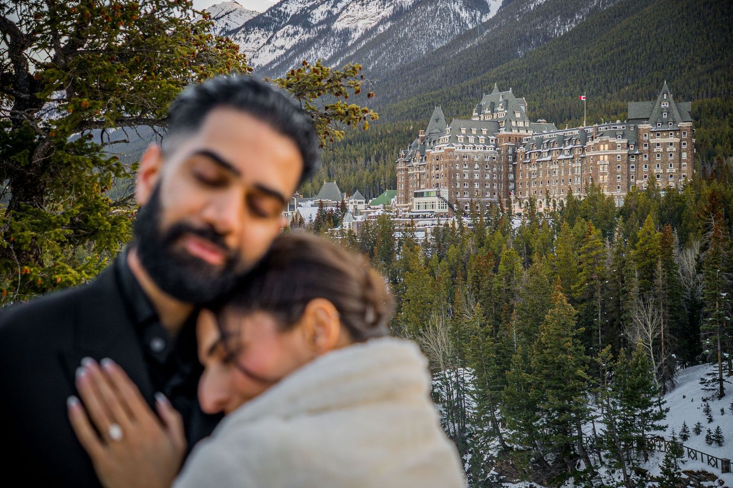 Romantic couple embraces in front of snowy mountain landscape with iconic Fairmont Banff Springs Hotel in background.