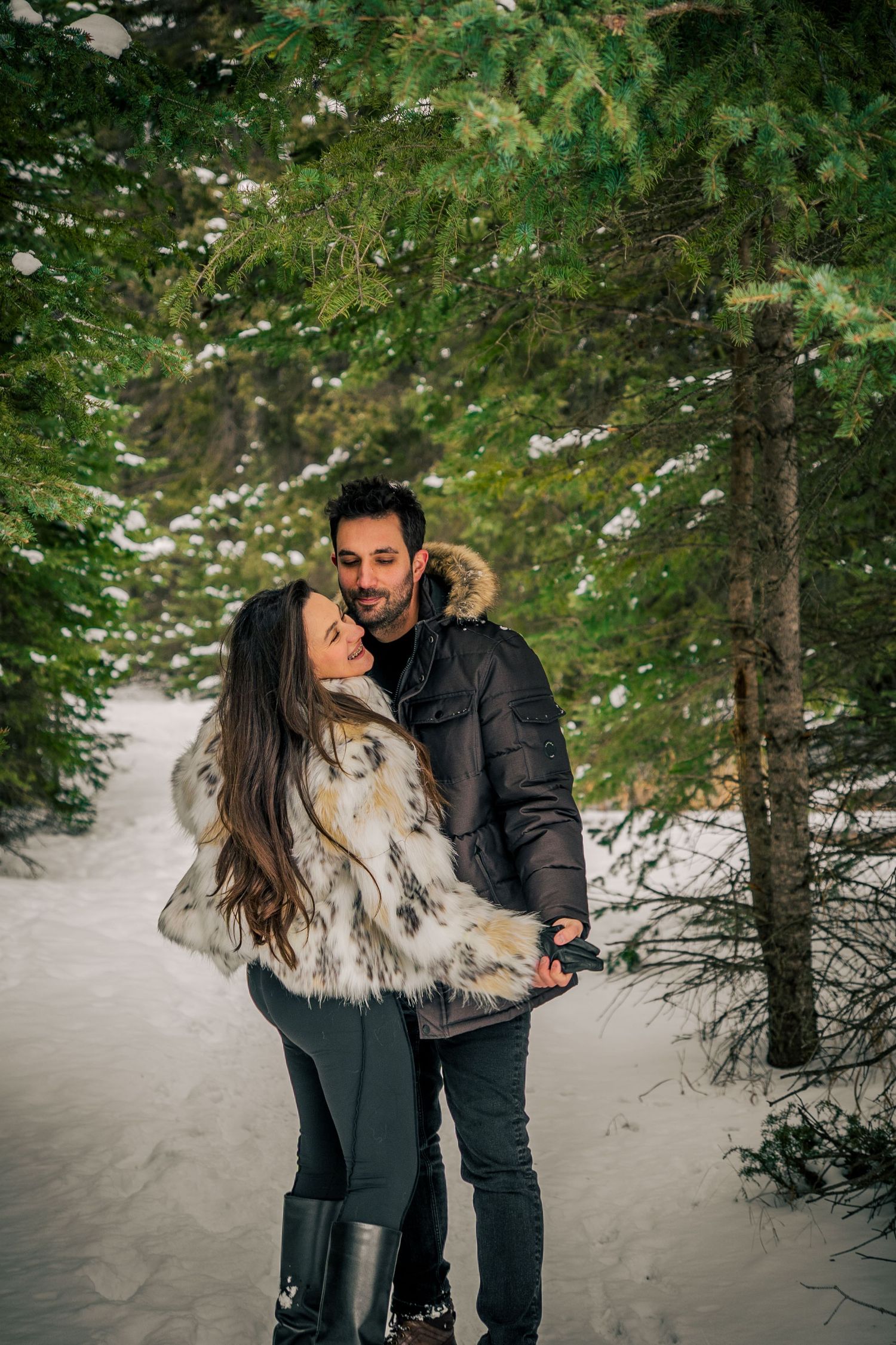 A couple shares a romantic moment in a snowy forest setting, wearing winter coats and embracing each other.