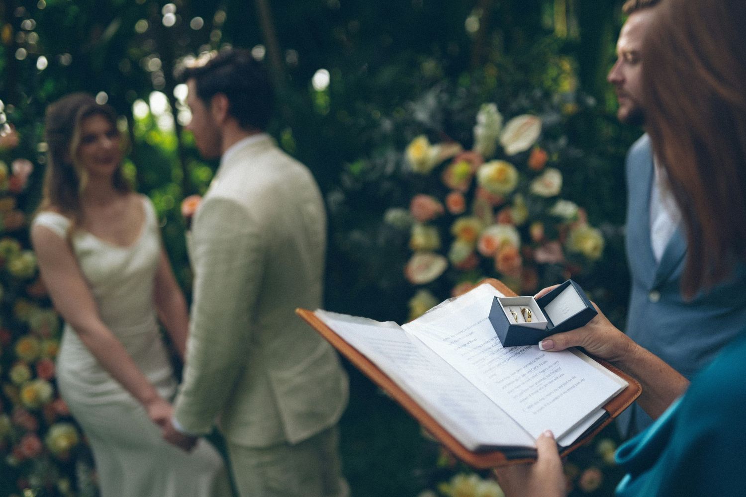 A person is holding an open book in front of a couple standing in a garden with flowers.
