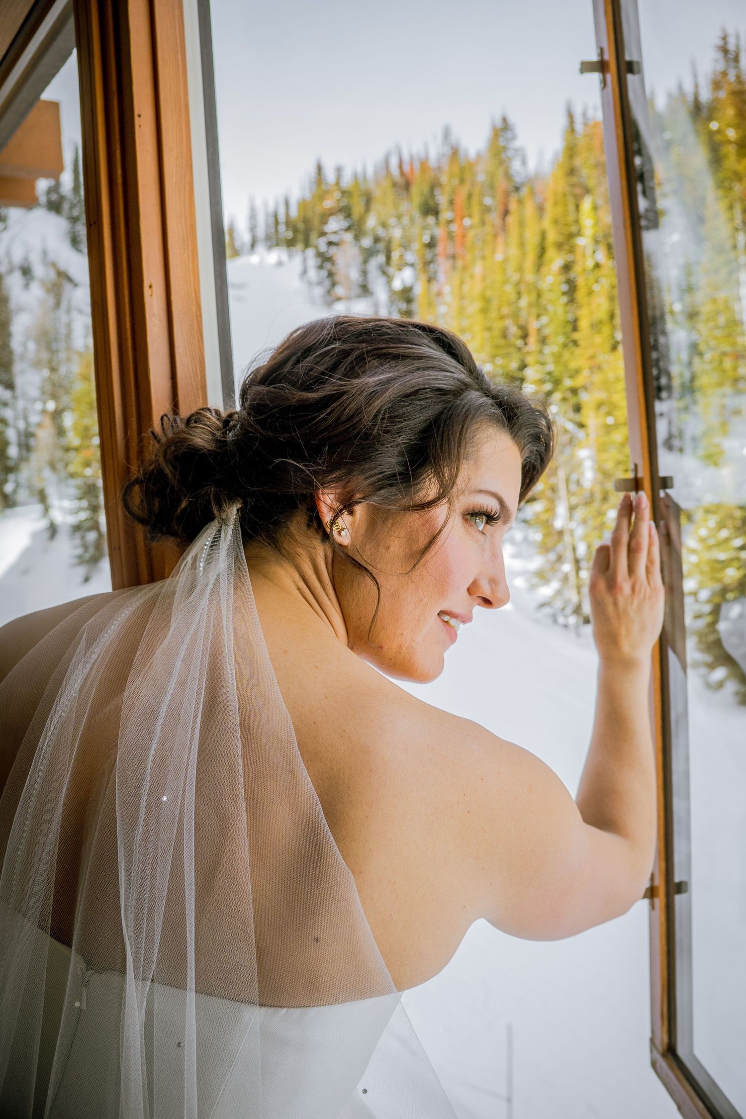 Bride in a white dress and veil looks out a window with snowy evergreen trees visible outside.
