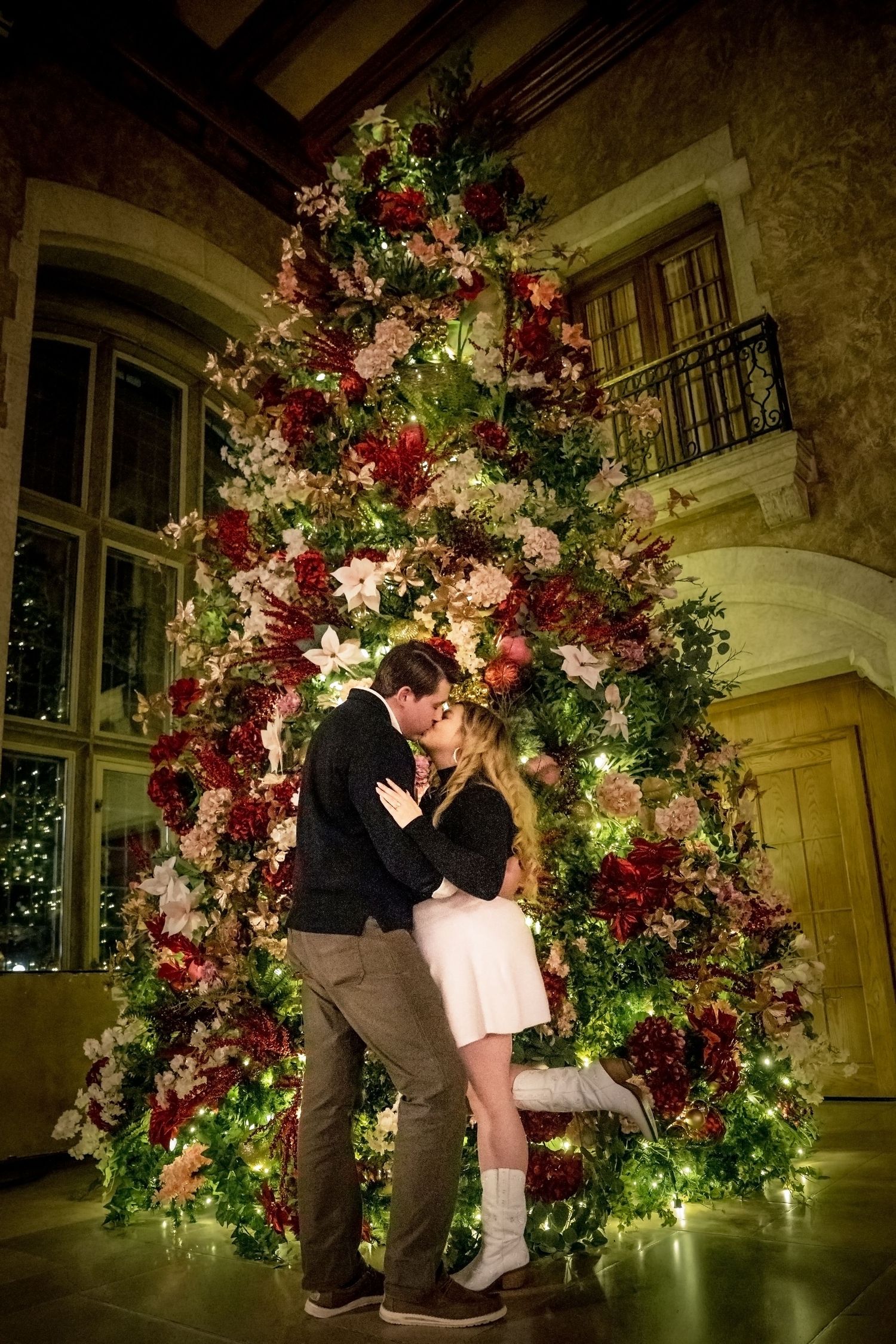 A romantic couple shares a kiss under an illuminated floral arch decorated with pink and white blooms.